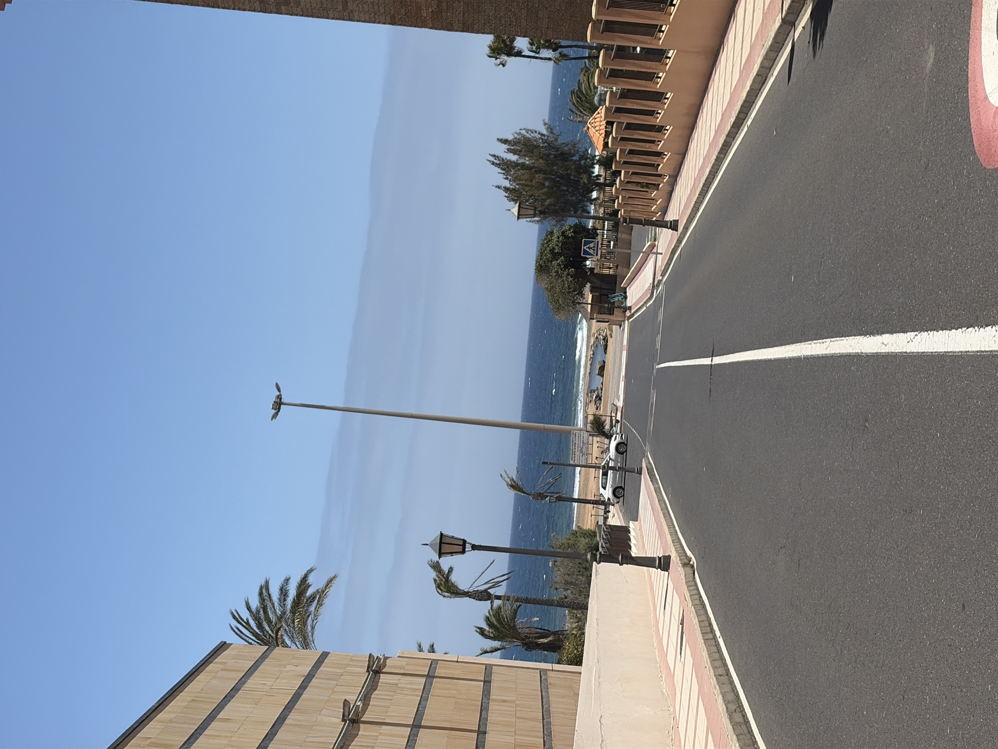 The image shows a street in Antigua, Spain, leading towards the ocean. On the left, there is a modern building with a textured beige facade and dark horizontal accents. Palm trees with fronds bent by the wind are visible. The street is paved with asphalt and has a white dividing line. There are sidewalks with a pattern of beige tiles and pinkish-red stones. A lamppost stands near the sidewalk. In the middle distance, a white car is parked on the side of the road. Beyond the car, the ocean is visible, with waves and a sandy beach. A tall, modern street lamp is positioned further down the road, and several other lampposts are scattered throughout the scene. On the right side of the road, there is a decorative fence made of dark metal and topped with terracotta-colored material. Further back, more trees and the ocean are visible. The sky is clear and blue, with some light clouds near the horizon. The overall impression is a sunny day in a coastal town.