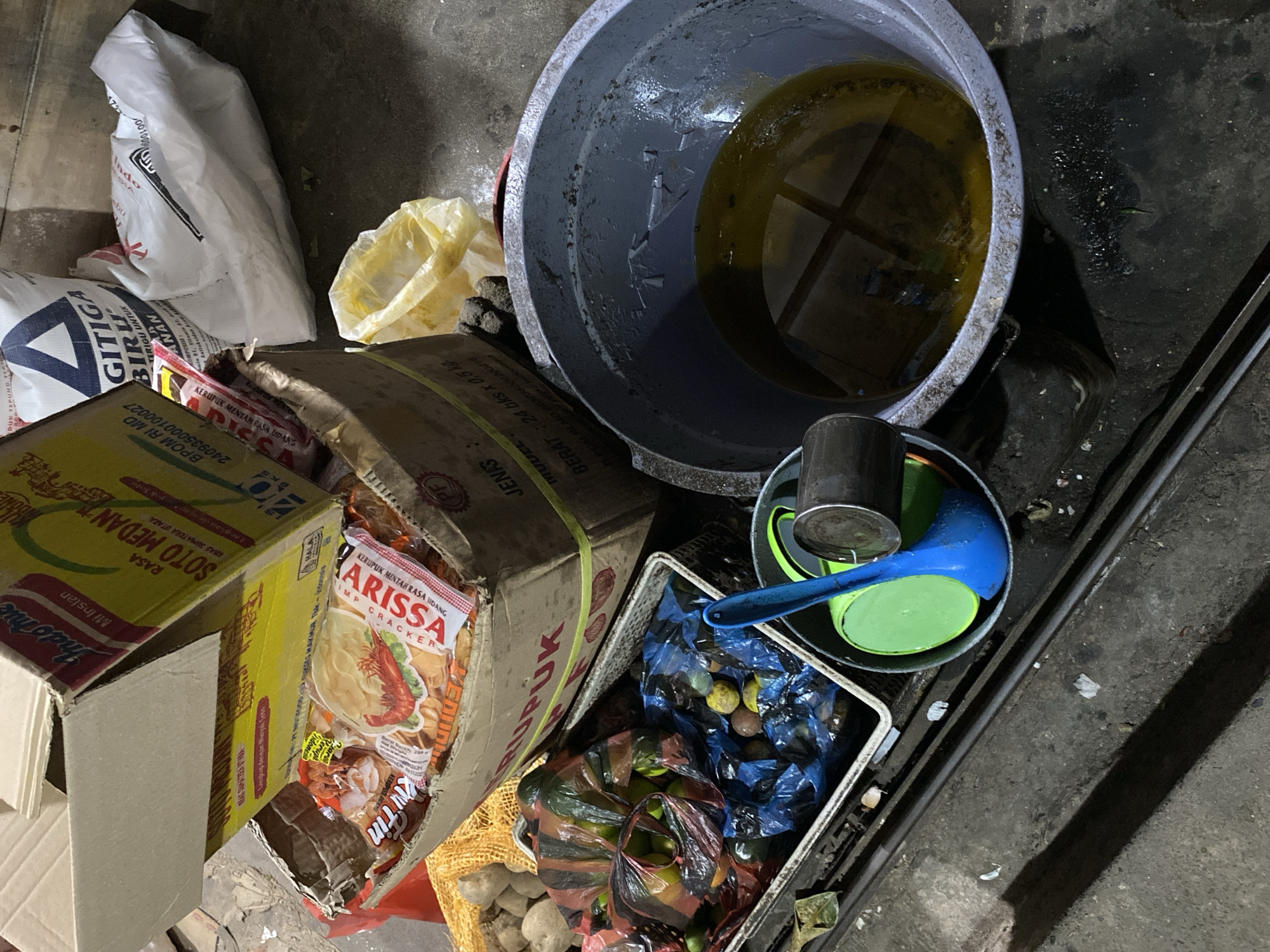 The image depicts a pile of food items and containers on a rough, concrete surface, potentially in a market or storage area. In the foreground, a large, gray, weathered bucket contains a yellow, oily liquid. To the left of the bucket is a white plastic bin containing what appears to be vegetables or fruits, some wrapped in colored plastic bags. Behind this bin, a cardboard box is tilted open, revealing packets of instant noodles and other food packaging, some featuring images of shrimp and text in Indonesian. Further back, a large white sack with blue lettering is visible, along with another yellow plastic bag. The overall impression is one of a disorganized collection of goods, possibly awaiting sale or disposal, in a dimly lit, utilitarian environment. The specific location within Aneuk Paya, Indonesia, cannot be definitively determined from the visual evidence alone, as there are no identifying landmarks or signs.