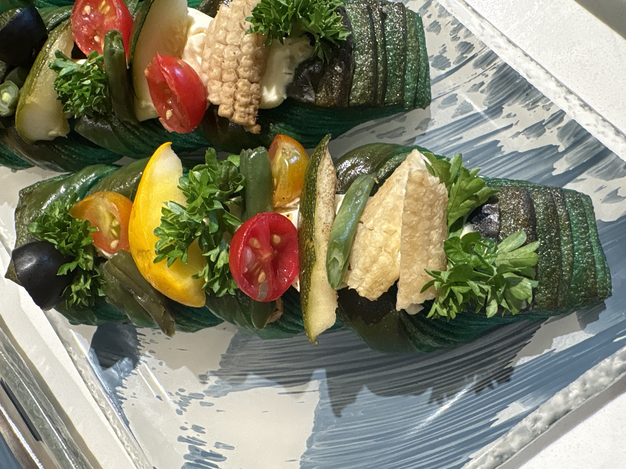 This is a close-up, overhead shot of two appetizers arranged vertically on a textured, light-colored serving platter with blue brushstrokes. Each appetizer is a layered creation topped with various vegetables and garnishes. The appetizer on the left features segments of green beans, cherry tomatoes (red and yellow), a slice of yellow bell pepper, and a dark olive. The appetizer on the right is topped with a slice of corn on the cob, a dollop of white sauce, and chopped parsley. Both appetizers appear to be wrapped in dark green, possibly grilled or roasted vegetable layers, with a glittery texture. The overall presentation suggests a sophisticated culinary offering. The location is not discernable from the image.