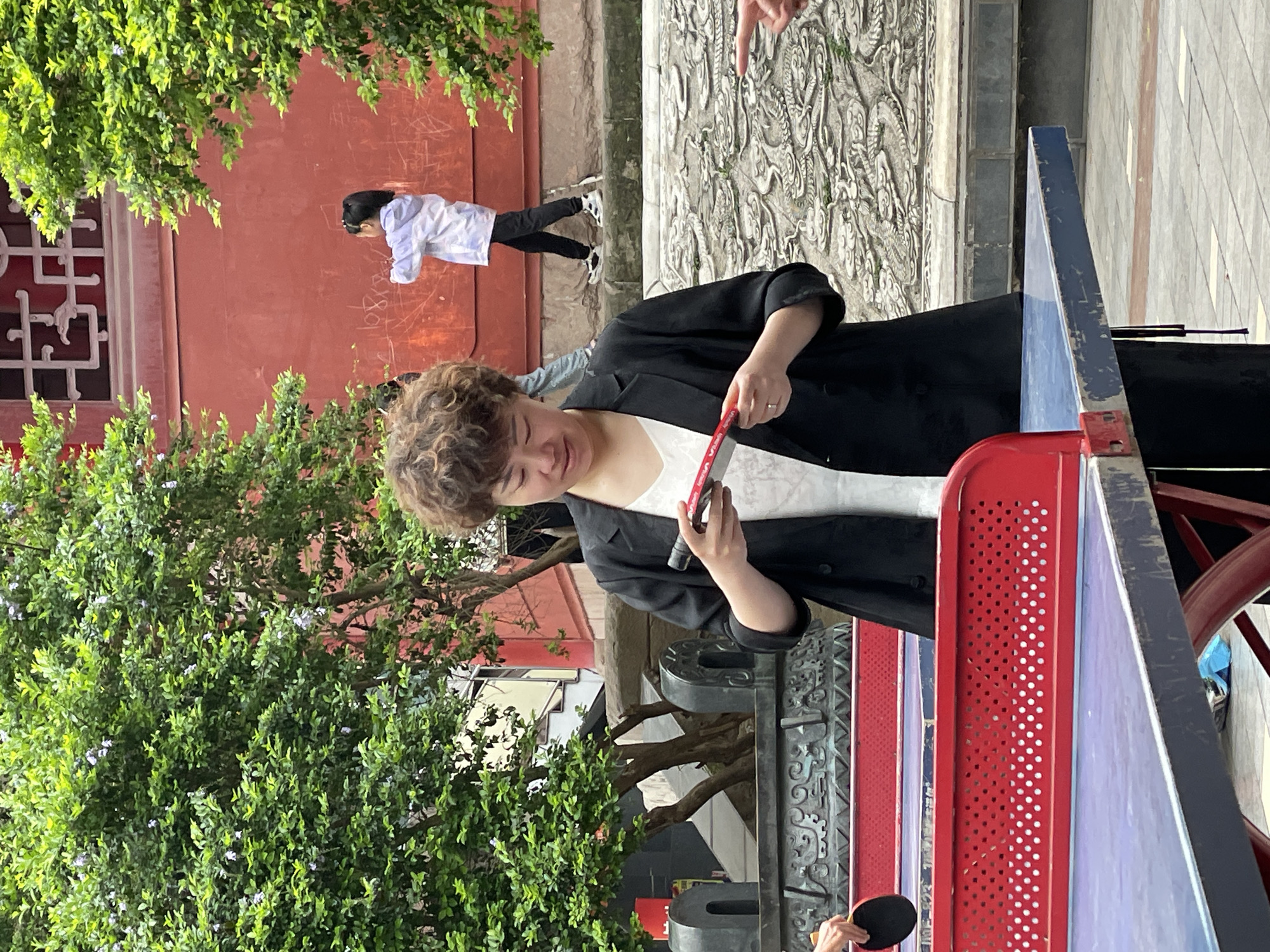 A person with curly hair, wearing a white shirt and a black jacket, is holding a red and black ping pong paddle. They are standing near a ping pong table with red and blue surfaces. A second person, wearing a white shirt and black pants, is writing on a red wall in the background. A large tree with green leaves is in the foreground on the left side of the image. Behind the person with curly hair is a stone wall with intricate carvings. The scene appears to be outdoors in the city of Jiangjin, China.
