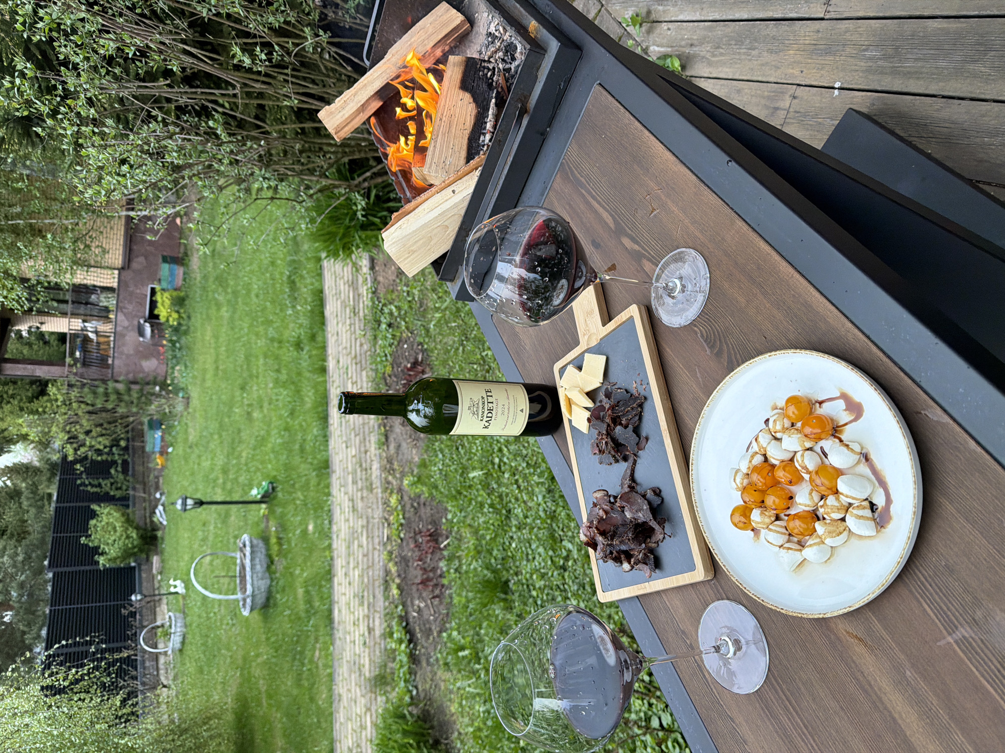This image shows a table setup outdoors, likely in a backyard setting. A fire pit containing burning logs sits at the edge of the table. On the table are two wine glasses filled with red wine, a bottle of red wine, a charcuterie board with cheese and biltong, and a plate containing what appears to be mozzarella balls and cherry tomatoes drizzled with balsamic glaze. The background features a green lawn with various decorative elements such as metal baskets and a garden light. The scene suggests a relaxing outdoor dining experience.
It is not possible to confirm the location of this scene as Zimyonki (Zimenki, Zimenka), Russia based on the image.