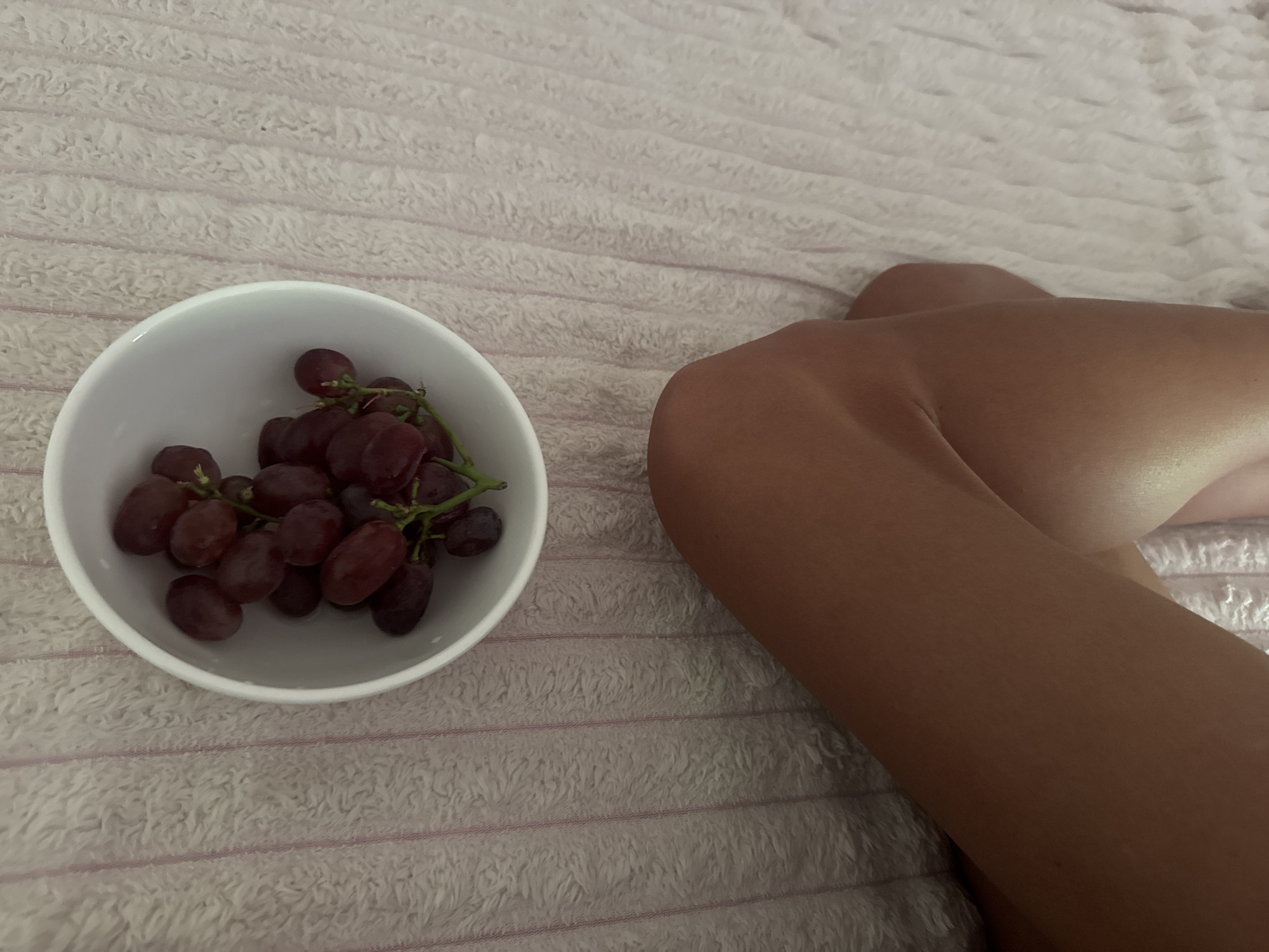 A top-down, slightly angled shot shows a white bowl filled with red grapes. The bowl is positioned in the upper center of the frame. Below the bowl, the lower half of the image features a person's tanned legs, bent at the knees, positioned diagonally from the lower left to the mid-right. The background is a textured, pale pink surface with vertical stripes. The lighting appears to be natural and soft.