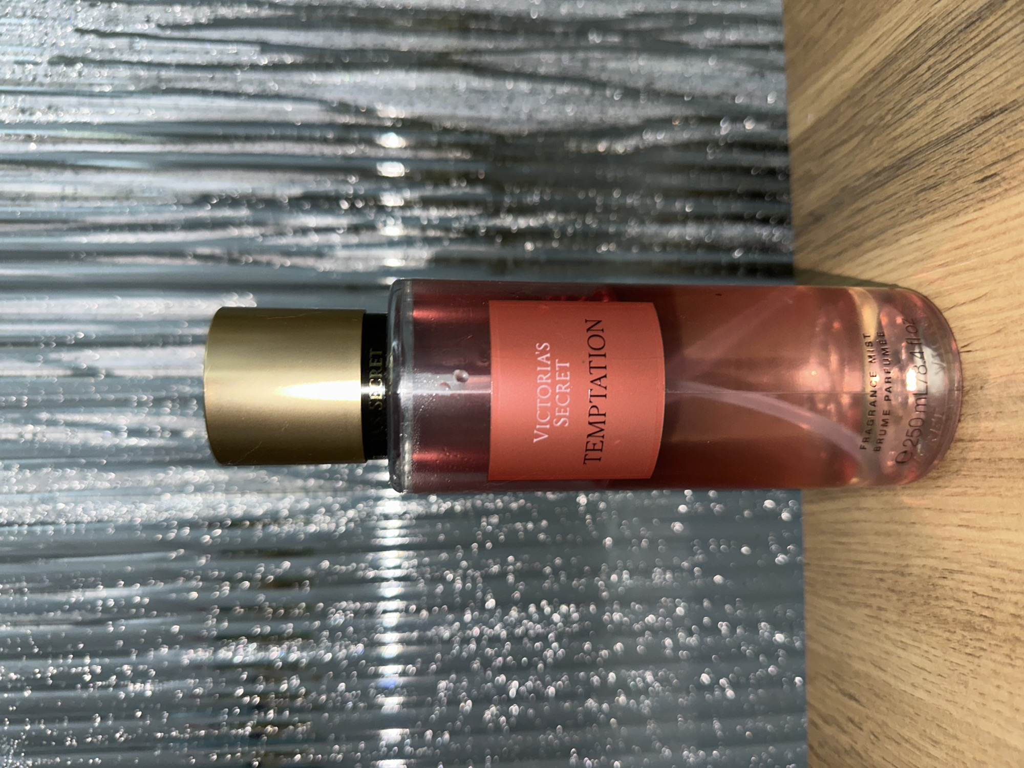 The image shows a bottle of Victoria's Secret Temptation fragrance mist lying horizontally. The bottle has a gold cap and a pink label with the brand and scent name printed on it. The background is split between a silver, reflective surface and a light-colored wooden surface. There is no indication of the location in Chaikovsky, Russia, or any people present in the scene.