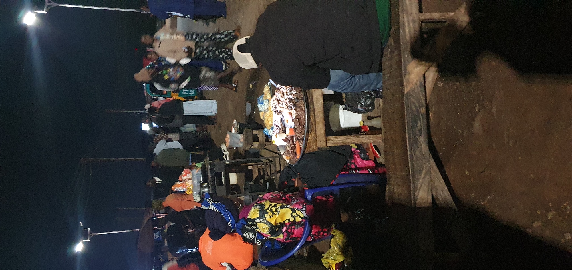 This is a nighttime street scene in Bukuru, Nigeria, depicting a bustling market with numerous people and food stalls. The lighting is provided by streetlights, casting a somewhat harsh glow on the scene. In the foreground, a man wearing a black jacket and a white baseball cap sits facing away from the camera, in front of a large circular metal tray filled with cooked food, possibly meat or skewers, along with some orange vegetables. To his left, a blue plastic chair is occupied by a person in a vibrant, patterned headscarf and a colorful, patterned dress. Next to the chair, a yellow plastic bag is visible.
Further back, several food stalls are set up. One stall features a metal grate over a fire, with pots and pans on it. Various people are gathered around these stalls, some appearing to be vendors and others customers. They are dressed in a mix of casual and traditional attire. In the background, a three-wheeled vehicle, common in Nigeria, is visible, along with more stalls and a crowd of people. The ground appears to be dirt or unpaved road. The overall impression is one of lively nighttime commerce, with food being prepared and sold to a diverse group of individuals.