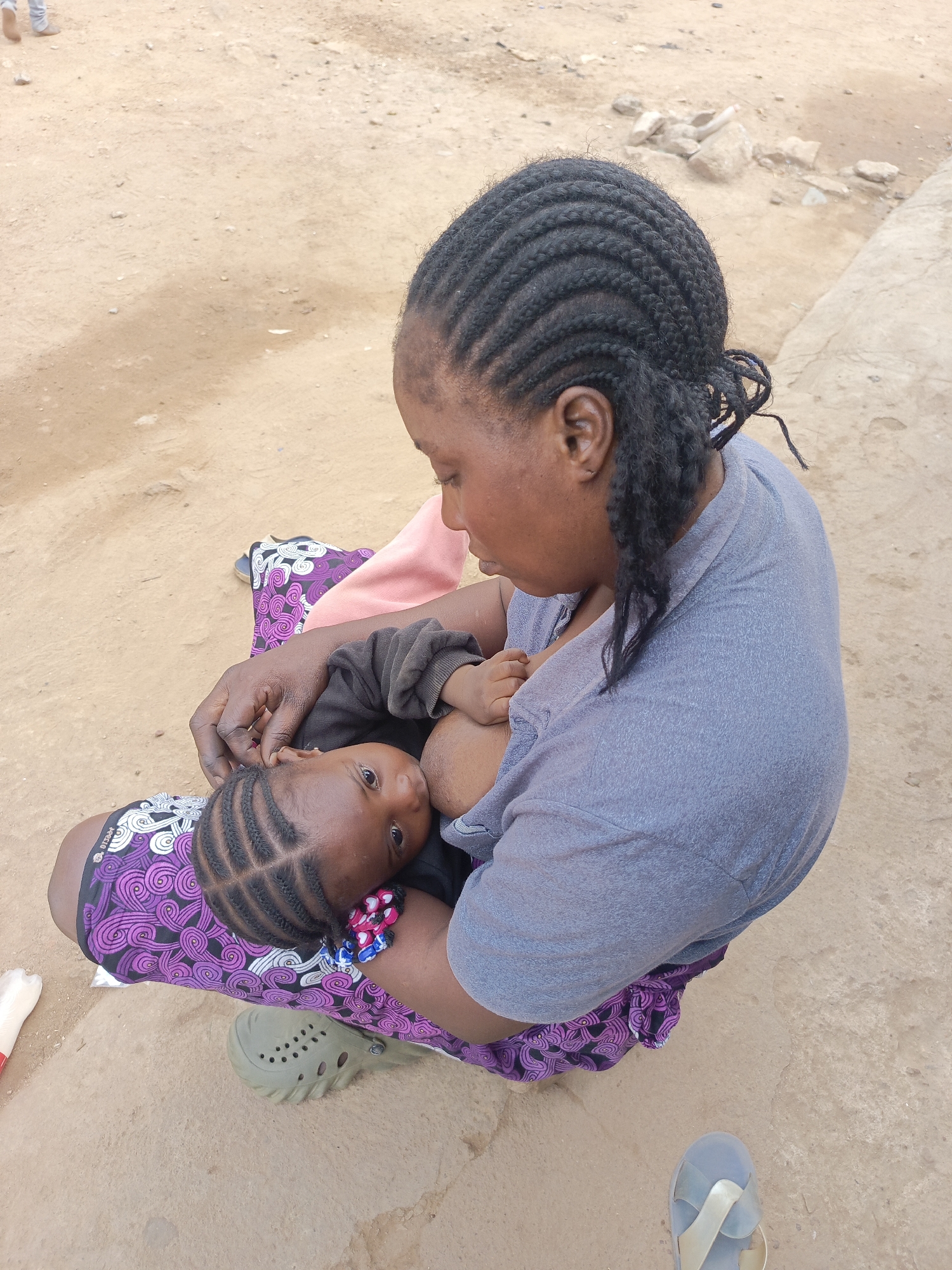 A woman with braided hair is breastfeeding a baby with braided hair. The woman is wearing a gray shirt and a purple and white patterned skirt. The baby is wearing a black shirt. The woman is sitting on the ground, and the baby is in her lap. They are in Akwanga, Nigeria. The ground is dirt. A pair of green Crocs and a pair of blue flip-flops are nearby.