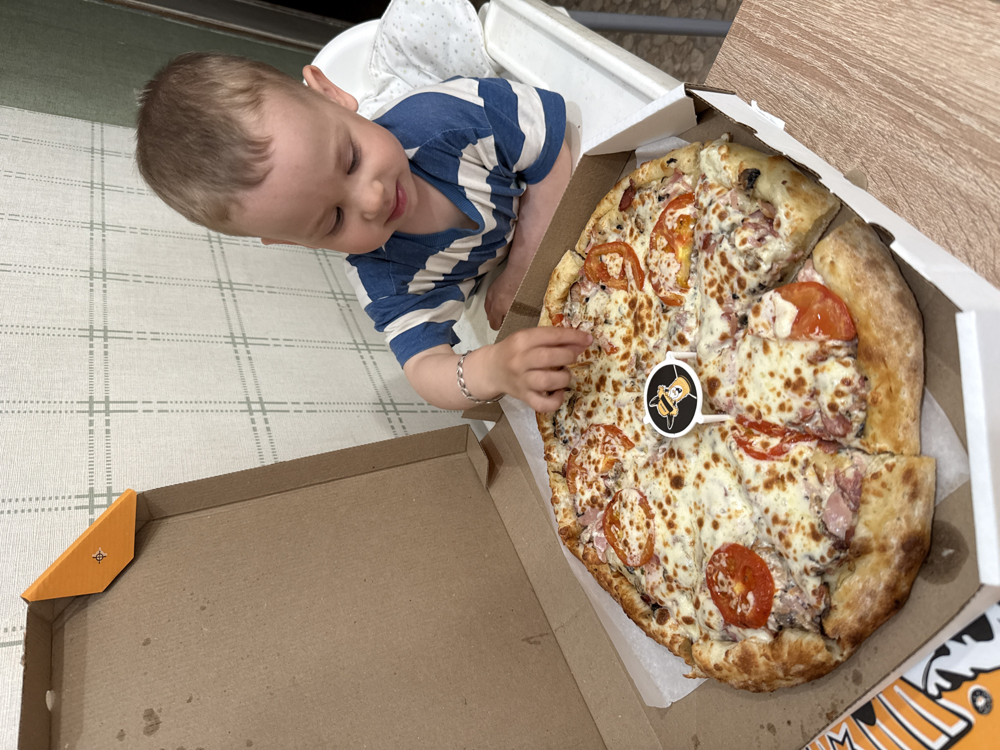 A young child in a blue and white striped shirt sits in a high chair and reaches for a pizza in an open pizza box. The pizza appears to be topped with tomatoes, ham, and cheese, and has a small logo stick in the center. The location is Заволжье, Russia. The pizza box is situated on a table next to the child. A second section of the pizza box lies open to the left of the pizza, revealing its cardboard interior. The flooring beneath the high chair has a grid pattern. The tabletop is a light wood color.