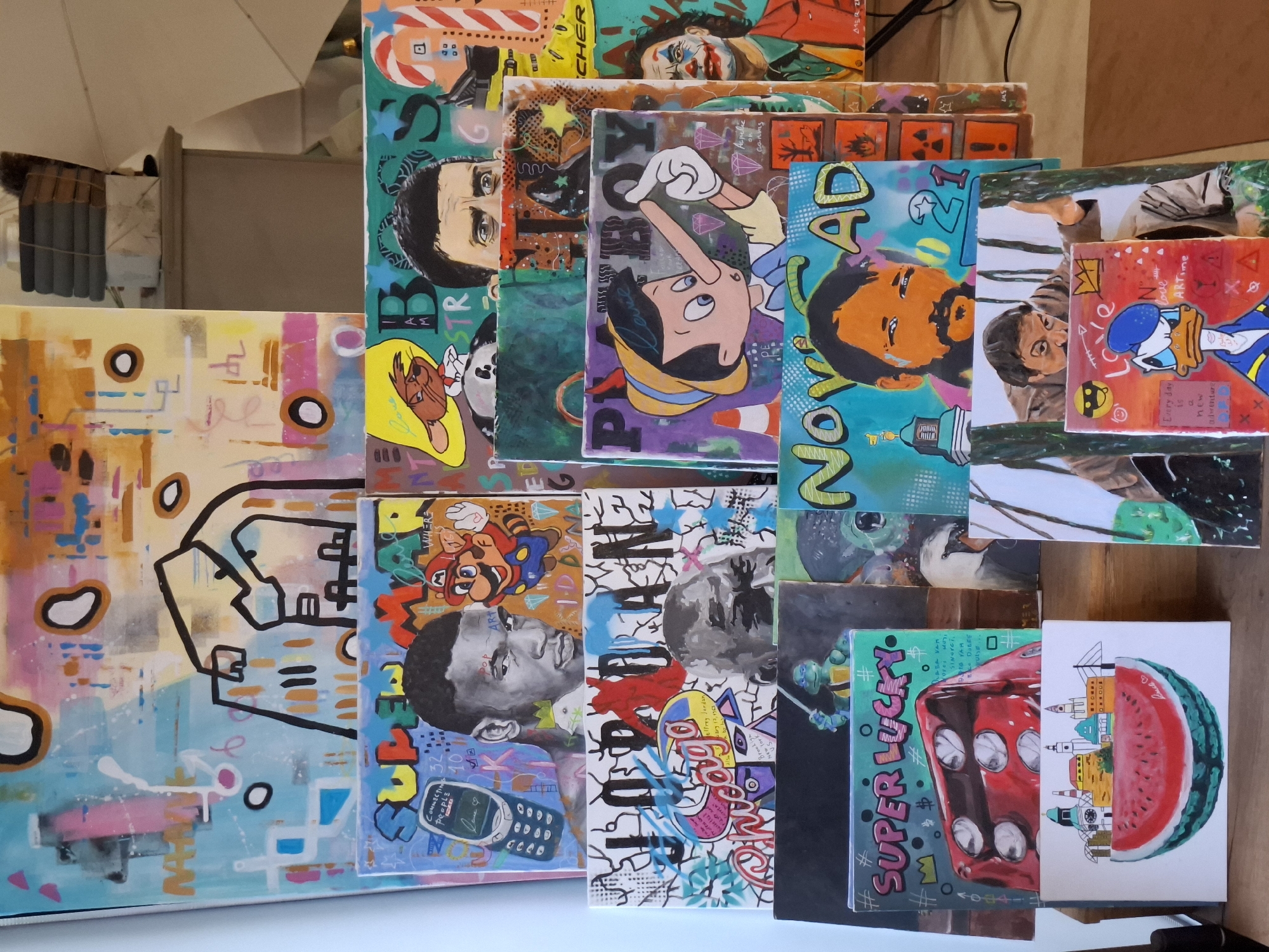 The image displays a collection of small, colorful canvas paintings stacked together, creating a collage-like composition. The paintings feature diverse subjects and styles, including portraits of people (e.g., Joker, Barack Obama), cartoon characters (e.g., Pinocchio, Donald Duck, Mario), abstract designs, and objects like a dice and an old cellphone.
One painting is of a watermelon slice, on which a skyline is drawn. This skyline is based on Novi Sad, Serbia.
The environment is indoors, possibly an artist's studio, and the lighting is somewhat diffused.
