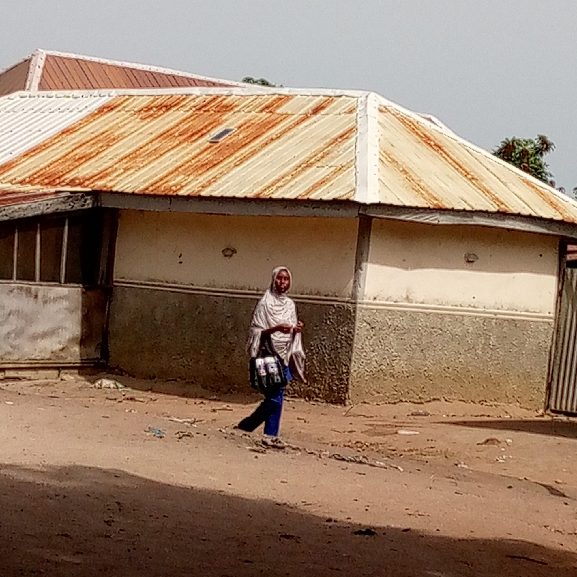 The image captures a street scene in Mararaba, Nigeria, featuring a lone woman walking past a building. The primary subject is a woman, seen from a medium distance, wearing a light-colored head covering and draped garment over dark trousers. She carries a dark shoulder bag with indistinct colorful patterns or text on her left side. Her gaze is directed slightly towards the camera as she walks across the unpaved, dusty ground.
The background is dominated by a single-story building with a distinctive, multi-sloped corrugated metal roof. The roof is significantly rusted, displaying a mix of orange-brown and lighter, possibly silver, patches. A small, dark rectangular object, possibly a solar panel or vent, is visible on one section of the roof. The building's walls are two-toned: the upper section is a light, creamy white, while the lower section is a darker, textured gray-brown. On the left side of the building, several dark-framed window-like openings are visible. A corrugated metal gate is partially seen on the far right.
The ground is an uneven dirt road with scattered small debris. Shadows in the foreground suggest a light source from the side or behind the photographer. The sky is a uniform, light gray, indicating either an overcast day or bright, diffuse daylight. The overall impression is that of a typical residential or commercial area in an unpaved urban or semi-urban setting. No clear text is legible on the woman's bag due to the resolution.
