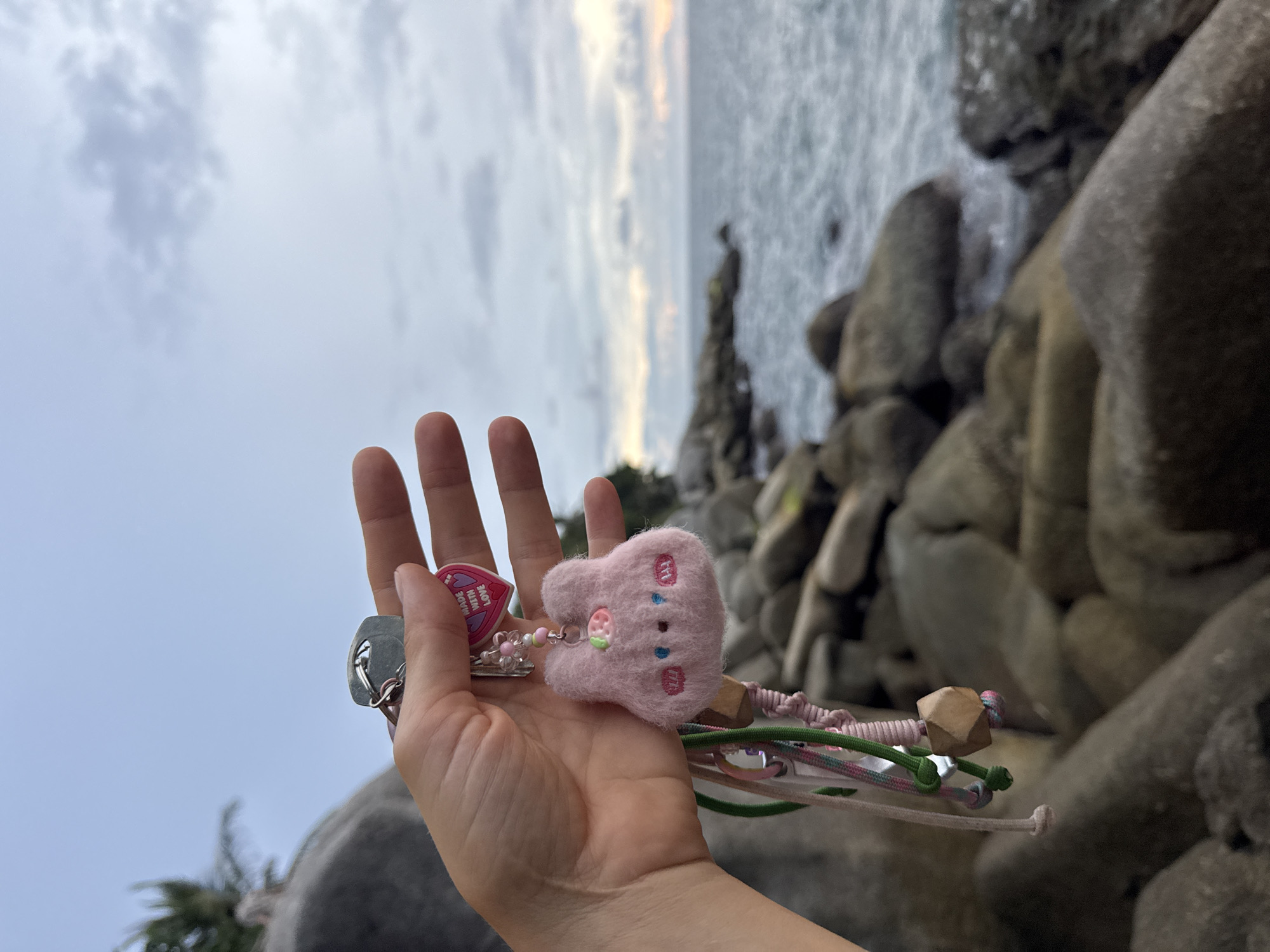 In Karon, Thailand, a hand holds a set of keys attached to a pink, fluffy bunny keychain with embroidered facial features and a small strawberry accessory. The keychain also features a small pink heart charm with the text "MADE WITH LOVE". The background shows a rocky coastline with the ocean and a cloudy sky.