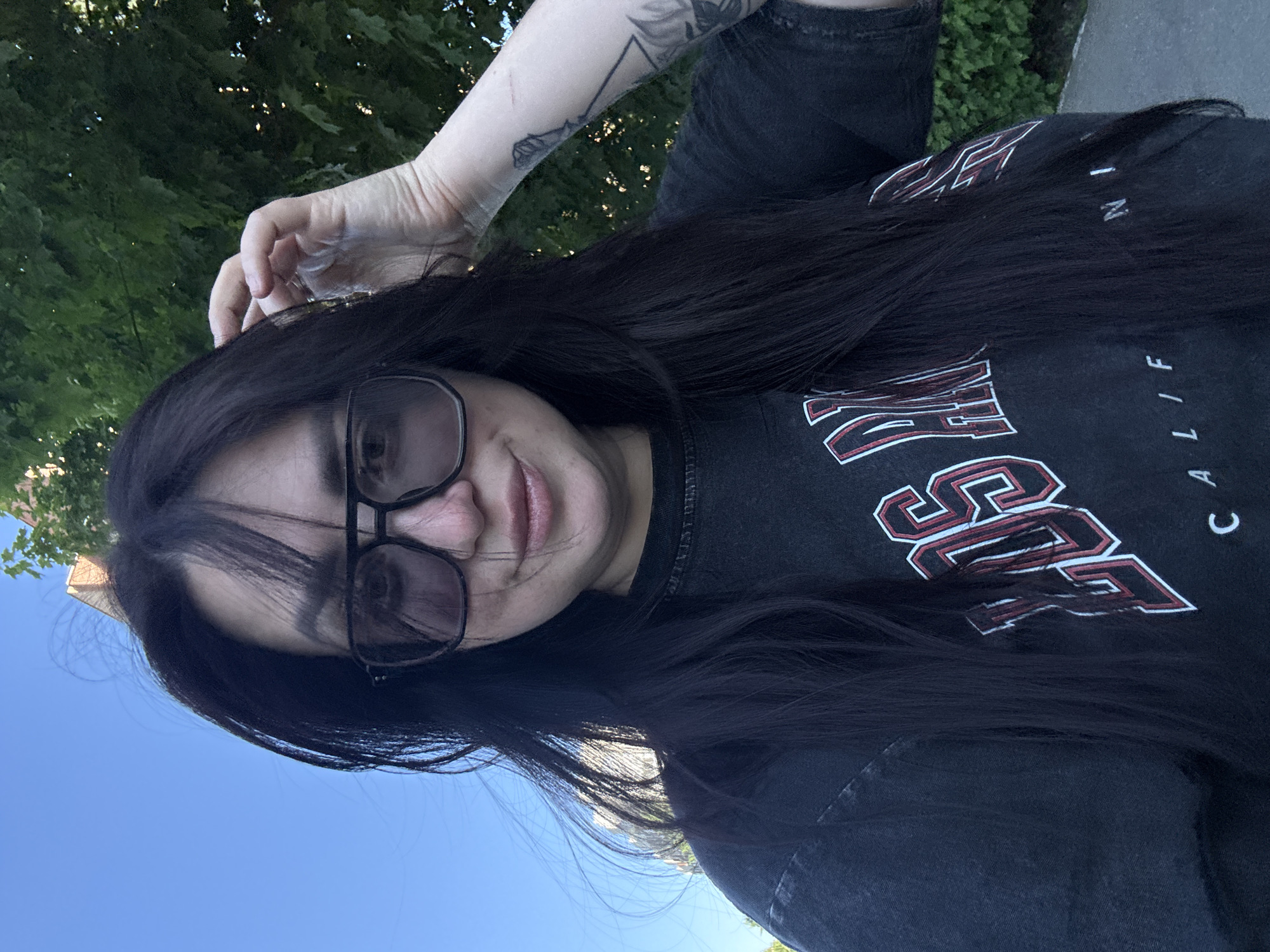 A woman with long, dark hair and black-framed glasses is the subject of this selfie, taken outdoors. She is wearing a dark-colored t-shirt with white and red lettering, with a visible tattoo on her left arm. The background shows trees and a clear blue sky, suggesting an urban park setting. The location in the city of Unknown, Unknown.