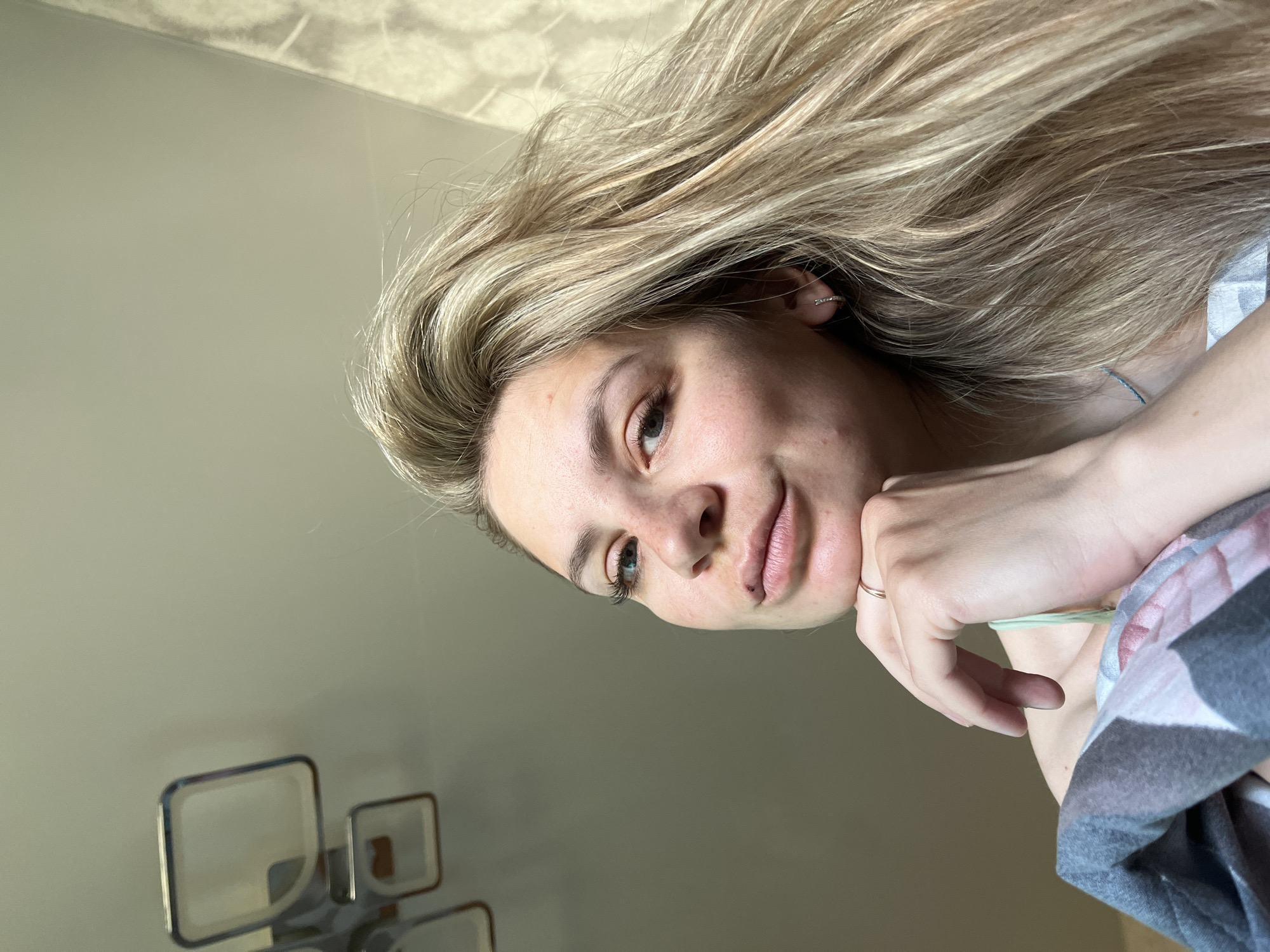A medium close-up, rotated, portrait of a blonde-haired woman is presented, resting her chin on her hand with a gold ring, revealing a green rubber band around her wrist. Her fair complexion is punctuated by light freckles. To the left of the frame are rectangular, chrome-accented wall decorations. The context of Chaikovsky, Russia, is not directly evident from the visual information.