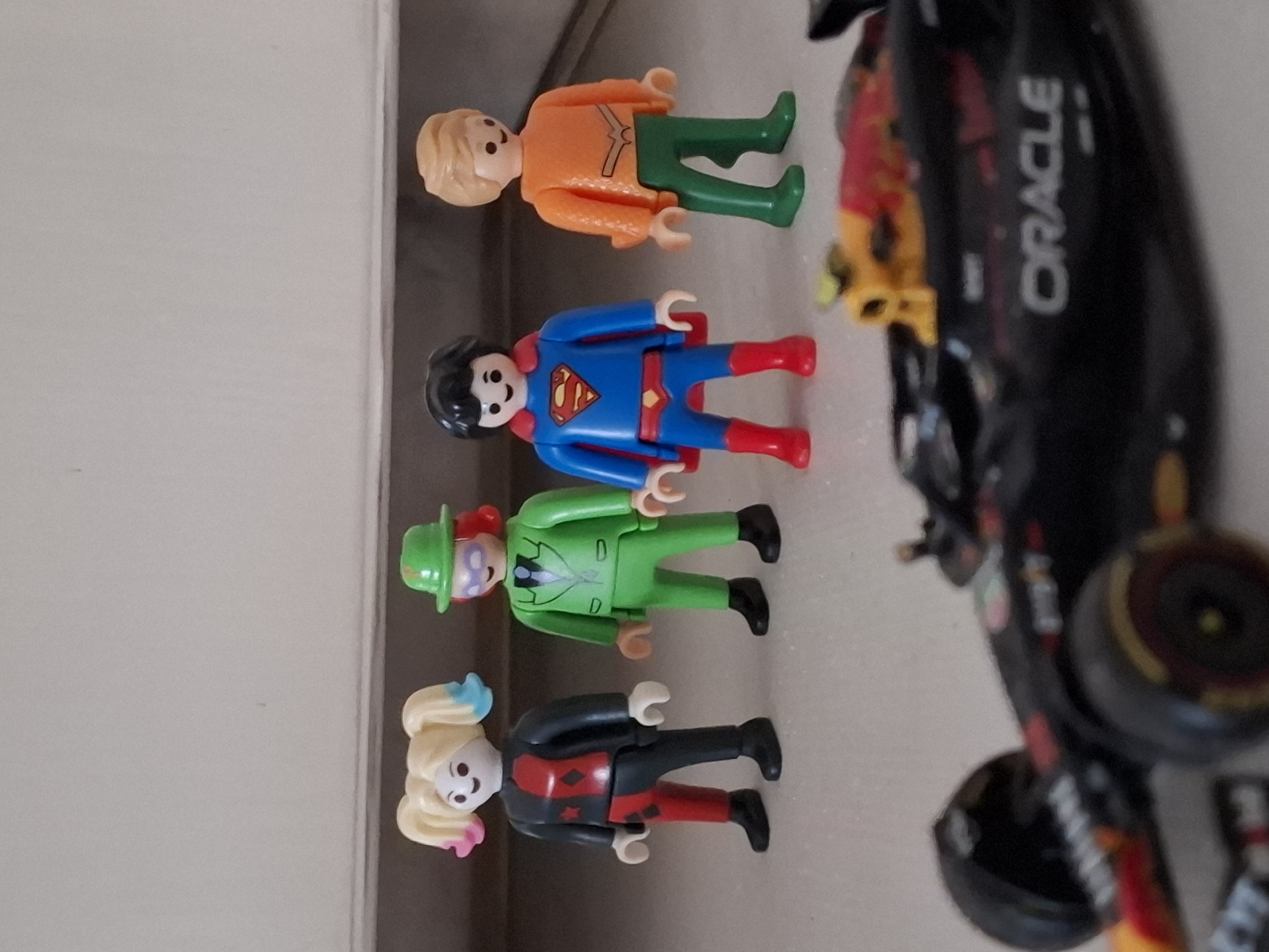 The image displays a collection of toy figures, likely Playmobil, arranged on a light-colored surface. From top to bottom, the figures include a person in an orange top and green pants, followed by a figure dressed as Superman in his iconic blue suit with red cape and boots. Next is a character wearing a green suit and hat, resembling the Riddler from Batman. The bottommost figure is a female character with blonde pigtails, dressed in a black, red, and white outfit, suggesting Harley Quinn. Partially visible on the right side of the image is a black toy race car, possibly a Formula 1 car, with "ORACLE" clearly visible in white text on its side. The overall scene appears to be an indoor setting, with soft lighting and a plain background, suggesting a display or play area. The toys are standing upright, implying they are posed for the photograph. There are no clear indicators of the time of day, weather, or specific location beyond the general context of a toy collection. The emotional tone is neutral, as expected from toy figurines.