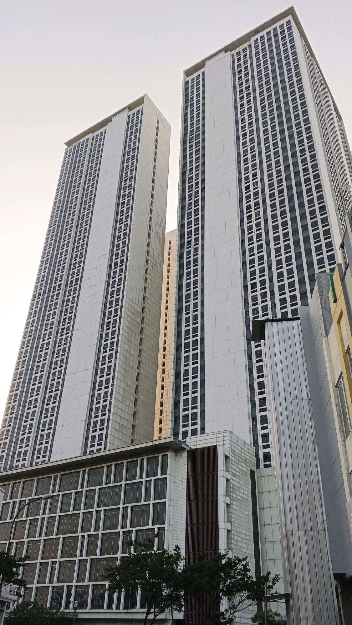 The image captures a low-angle view of two tall, modern residential buildings in Pasirsari, Indonesia. The buildings are predominantly white with numerous dark-colored windows arranged in a grid pattern, creating a repetitive and uniform facade. The building on the left is slightly shorter than the one on the right. Both structures feature sharp, angular designs. In the foreground, a lower, multi-story building with a glass-paneled facade is visible, adorned with vertical dark brown elements on its side. The lower part of the image shows the tops of dark green trees. The sky above is overcast and pale, suggesting either early morning or late afternoon light. No people are visible in the frame. The overall scene depicts an urban environment characterized by contemporary architecture.