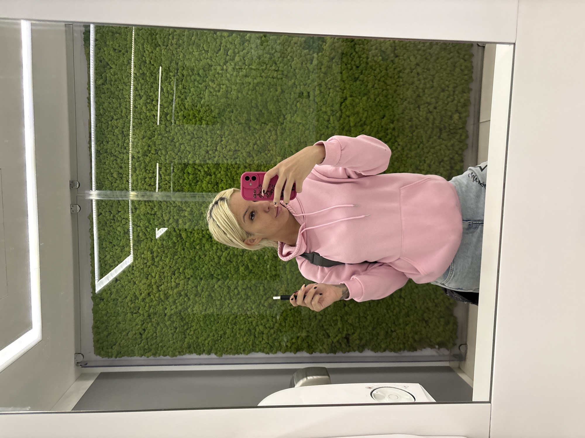 A photograph depicts a woman reflected in a mirror, apparently taking a selfie with a pink-cased mobile phone. She is wearing a pink hooded sweatshirt and light blue denim jeans. The background behind the woman in the reflection reveals a green, moss-like wall that stretches vertically. She is also holding a black, cylindrical object in her left hand. The lower part of the mirror reflects part of a white, possibly ceramic fixture, which might be a sink or toilet. The scene appears to be indoors, possibly in a public restroom or enclosed space with decorative moss wall installation. The location of the city is Unknown, Unknown.