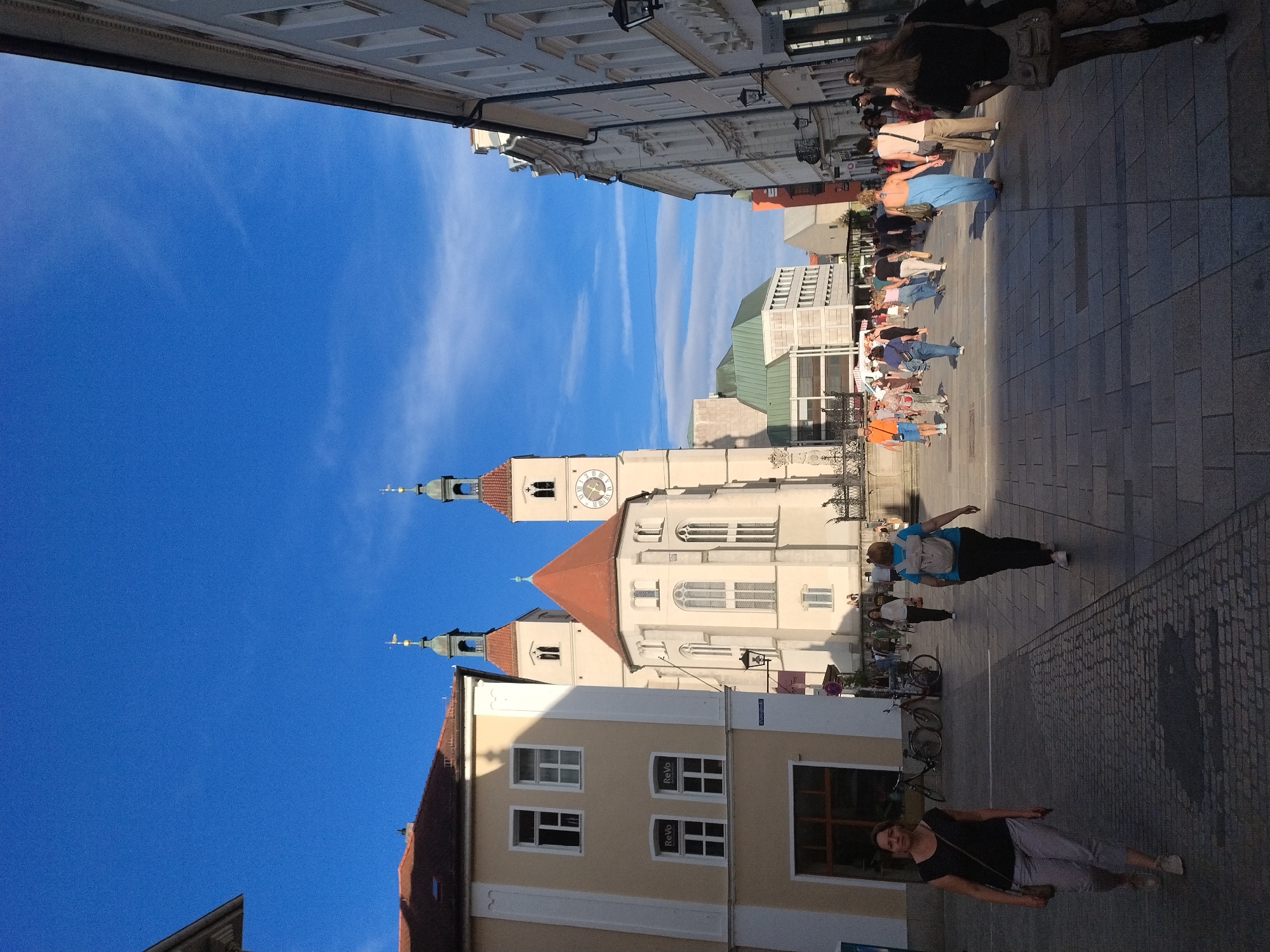 The image shows a view of the city of Regensburg, Germany, specifically looking towards St. Peter's Cathedral. The foreground features a cobblestone street with several people walking in various directions. To the left, a woman in a black shirt and gray pants walks towards the viewer. Further back, a woman wearing a blue backpack walks away from the viewer. The middle ground is dominated by the imposing Gothic architecture of St. Peter's Cathedral, with its two tall spires and a large clock on one of them. The cathedral is painted a light cream color and has a red-tiled roof. In front of the cathedral, there are more people walking and standing. The background consists of buildings lining the street, one of which is a light yellow building to the left of the cathedral. The sky above is a clear, bright blue with a few wispy clouds. The overall scene depicts a sunny day in a European city square with a prominent historical landmark.