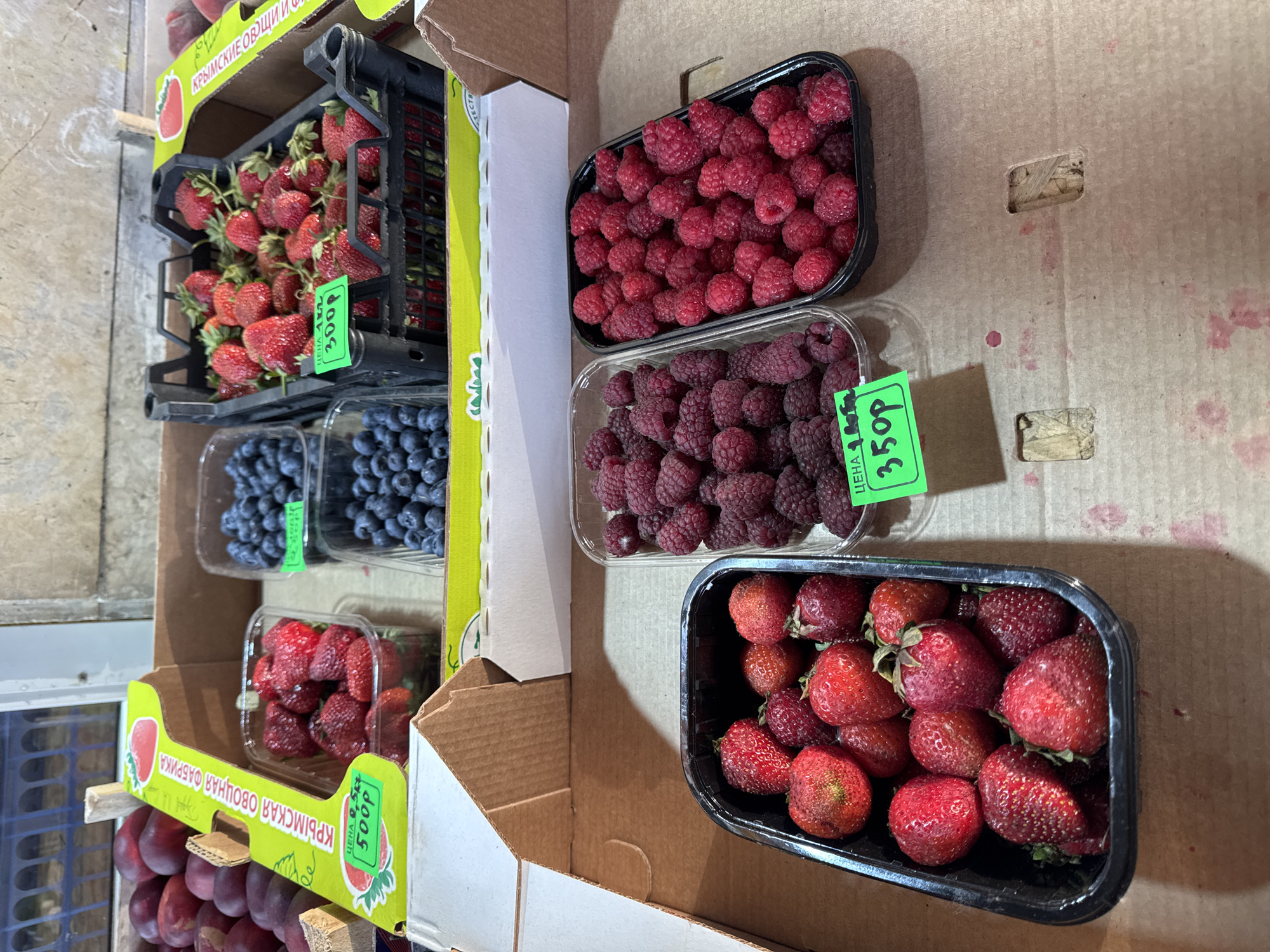 The image shows a close-up of a market stall in Saint Petersburg, Russia, displaying various fresh berries for sale. In the foreground, two black plastic containers are filled with ripe raspberries. A green price tag is visible on one of the raspberry containers, showing "350p". To the left, another black container holds a selection of strawberries. In the background, plastic containers of blueberries are visible, with one displaying a green price tag reading "300p". Further back, a bright yellow cardboard box with Russian text ("КРЫМСКАЯ ОВОЩНАЯ ФАБРИКА") contains more strawberries and what appear to be plums. The stall appears to be an outdoor or semi-outdoor market, with a wooden background and cardboard boxes serving as displays. The lighting suggests it might be daytime. No people are visible in the image.