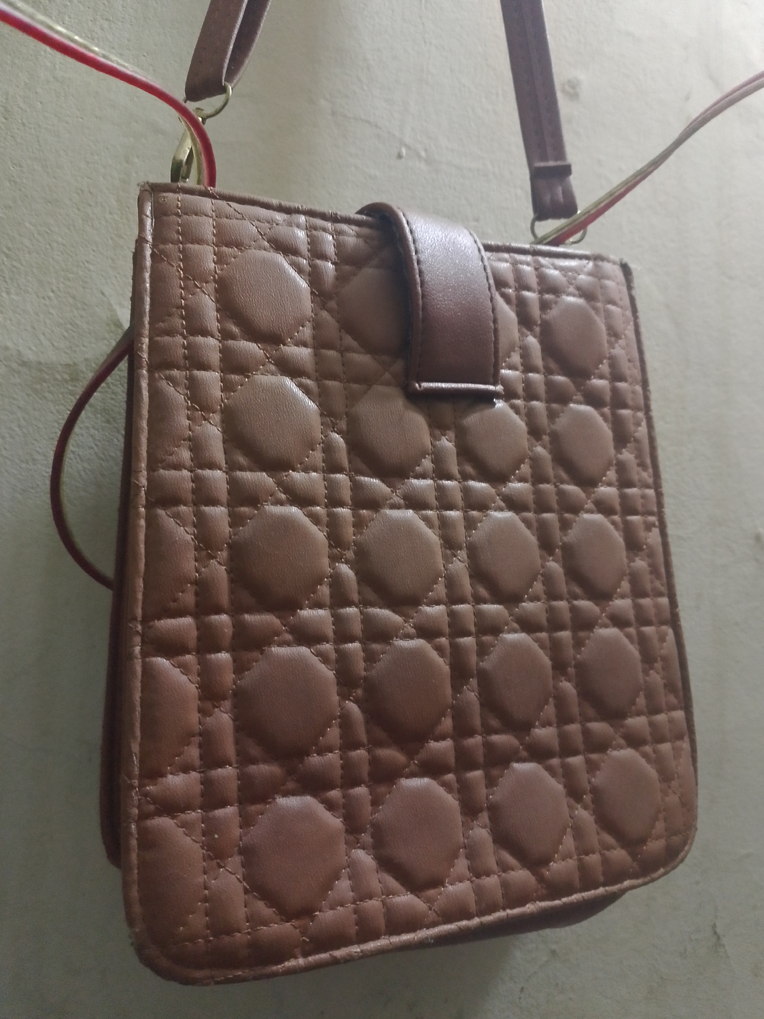 The image shows a brown, quilted, square-shaped handbag with a flap closure, hanging from a wall. The handbag has a pattern of diamond-shaped stitching with circular padded sections within each diamond. A thin, brown strap is attached to the top of the handbag, and two thin, red and green wires are also visible, attached to the strap. The background is a textured, light-colored wall. The image appears to be taken indoors. There is no indication of the specific location within Mukapayung, Indonesia.