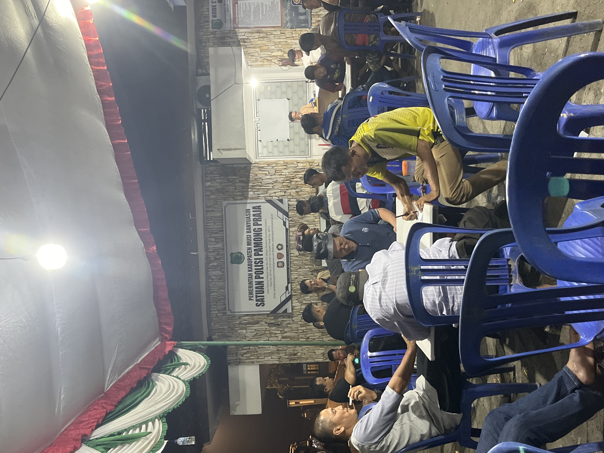 The image shows an outdoor gathering of approximately 20-25 individuals seated at tables under a tent. The setting appears to be in Sekayu, Indonesia, indicated by signage that reads "PEMERINTAH KABUPATEN MUSI BANYUASIN" and "SATUAN POLISI PAMONG PRAJA," with an address specifying "Jalan Kolonel Wahid Udin No. 237 Sekayu Provinsi, Sumatera Selatan." The individuals are predominantly adult males, dressed in casual attire, with some wearing uniforms or semi-formal shirts.
Several people are engaged in conversation, while others are looking at documents or writing. One person in the foreground is smoking a cigarette. The environment is dimly lit, with artificial lighting from a bulb overhead and some ambient light reflecting off a tent. The ground appears to be paved. Blue plastic chairs and white tables are arranged around the gathering space. In the background, there is a building with stone-patterned walls and a banner displaying official information. The overall atmosphere suggests a meeting, discussion, or informal gathering related to the local government or public service.