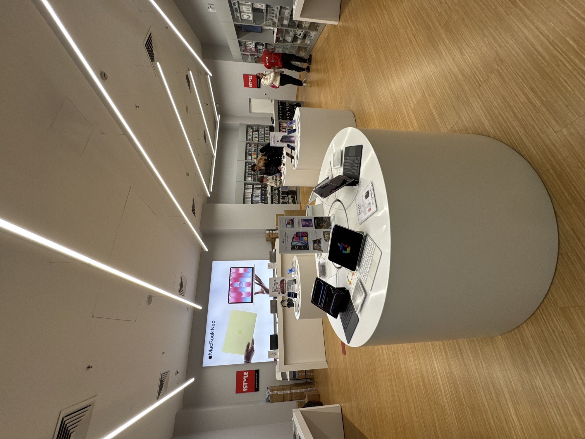 The image captures the interior of a modern electronics store, likely an authorized Apple reseller given the products and branding, located in Sofia, Bulgaria. The store features a clean, minimalist design with white walls, a light-colored wooden floor, and bright, linear strip lighting on the ceiling.
In the foreground and mid-ground, several white, cylindrical display tables are visible, showcasing various electronic devices. On the largest table in the foreground, multiple Apple iPads and MacBooks are arranged for display, including a black tablet with a keyboard case and a silver tablet with a white keyboard. Smaller display units and shelving line the walls further back, holding packaged products and accessories.
A large, illuminated wall display in the mid-ground prominently features a graphic with the text "MacBook Neo" and an image of a laptop. Below this, a red sign with the "iSTYLE" logo is visible. Another smaller "iSTYLE" logo is seen on a display stand towards the back.
Several people are present in the store, browsing the products. Two individuals are near the back wall, one wearing a red t-shirt and another in a striped top, appearing to look at items on the wall shelves. A group of a few more people are gathered around one of the central display tables.
The overall atmosphere is bright and inviting, typical of a retail environment during business hours. The store appears well-maintained and organized, with a focus on product presentation. Other notable details include air vents and small ceiling-mounted cameras or sensors.