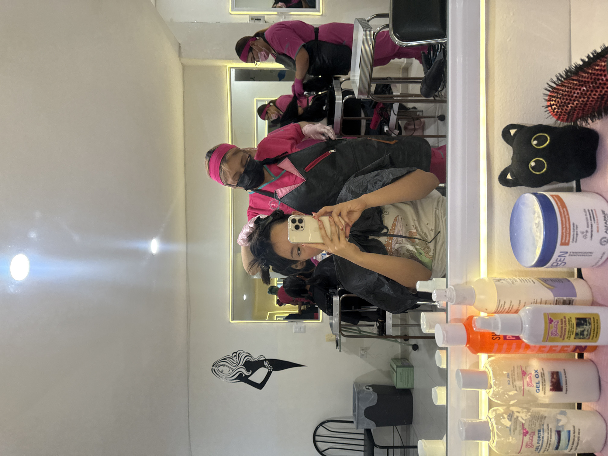 This image, taken in Puebla, Mexico, shows a woman taking a mirror selfie at a hair salon. The woman, who has dark hair pulled back and is wearing a black salon cape over a light-colored t-shirt with a design, holds a light-colored smartphone to capture her reflection.
In the background reflection, at least two salon staff members are visible. They are uniformly dressed in pink tops, pink headbands, and black aprons. One staff member, wearing a black face mask, appears to be actively working on a client's hair. Salon chairs and other equipment are also reflected in the mirror, indicating an active salon environment.
To the right of the mirror, a shelf holds various hair care products in bottles and jars. Visible text on these products includes "GEL FORTE," "GEL OX," "SP," "P," "SOLUCION PARA NUTRICION CAPILAR," "BEN," "ALFAPARF," and "galatea." A black cat-shaped plush toy with yellow eyes and a red and black hairbrush are also on the shelf.
The salon has white walls, with a decorative black silhouette of a woman's head and flowing hair on the wall to the left of the mirror. The space is well-lit by artificial lights, including fixtures on the ceiling and strip lighting around the mirror. The image, which is rotated 90 degrees clockwise, captures a casual moment during a salon visit.