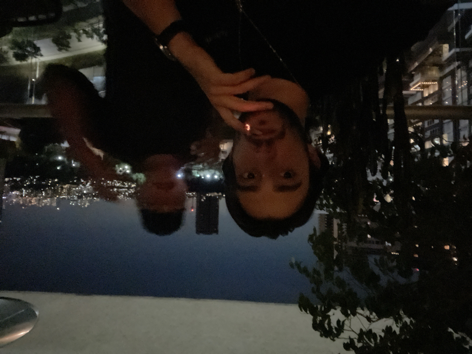 Two people are captured in an upside-down outdoor night shot, where the first person closer to the camera is holding a lit cigarette. The second person's face can be partially seen behind the first. The city of Huixquilucan de Degollado, Mexico is in the background, identifiable by the city lights in the distance. Foreground details include a low wall or ledge and dark foliage, suggesting an elevated viewpoint.
