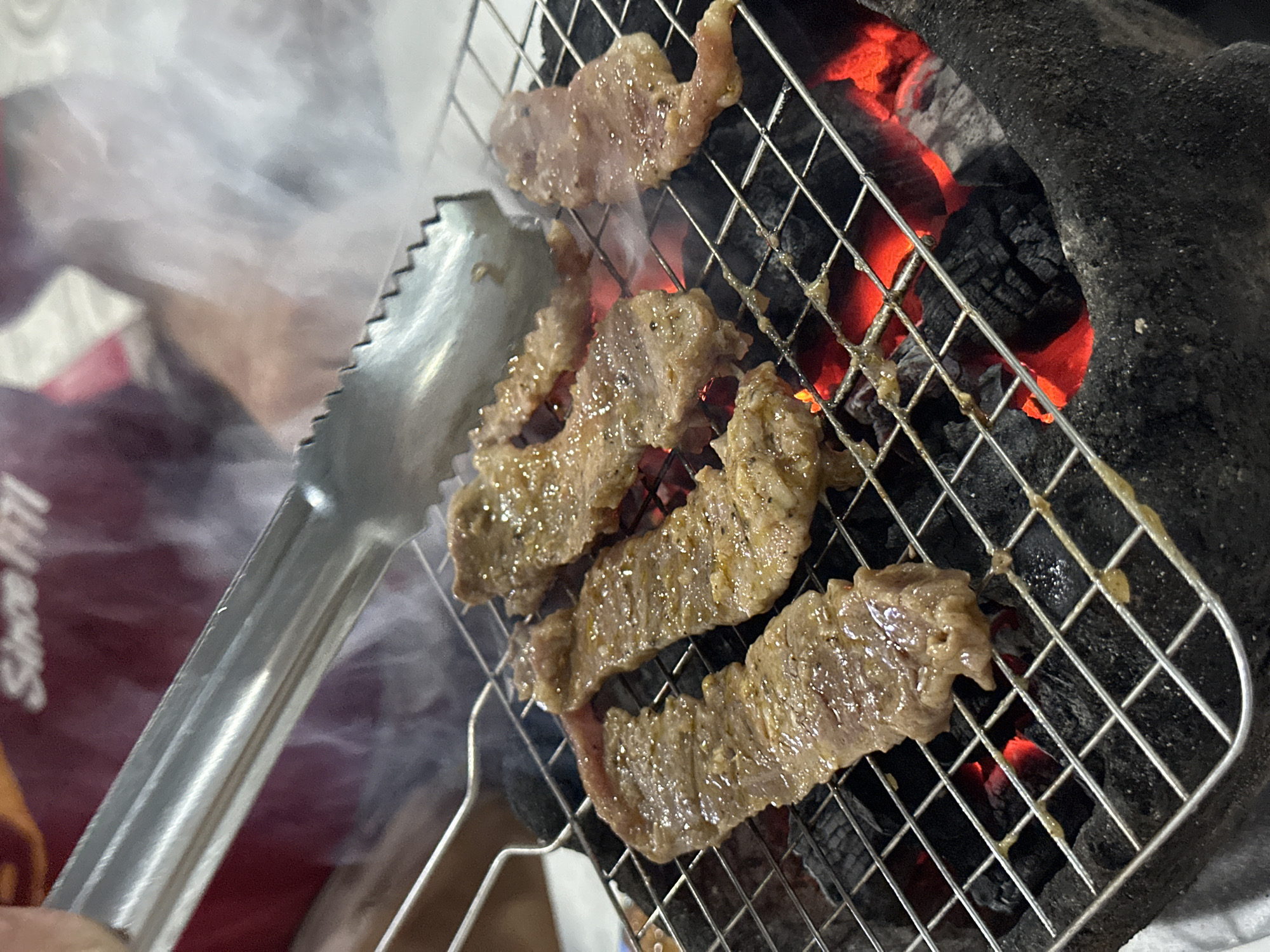 A close-up, low-angle photograph taken in Phu Thuy Ward, Vietnam, depicts thin slices of marinated meat being grilled over red-hot charcoal. The meat, possibly beef, is arranged on a metal grate above glowing orange-red charcoal embers within a dark, textured, possibly clay or concrete, brazier. Smoke rises from the sizzling meat. A silver-colored metal tong, held by an individual whose hand is partially visible on the left, is positioned above a piece of meat, indicating active cooking. In the blurred background, a person wearing a red garment is visible, with the white text "Since 1971" partially legible on the fabric.