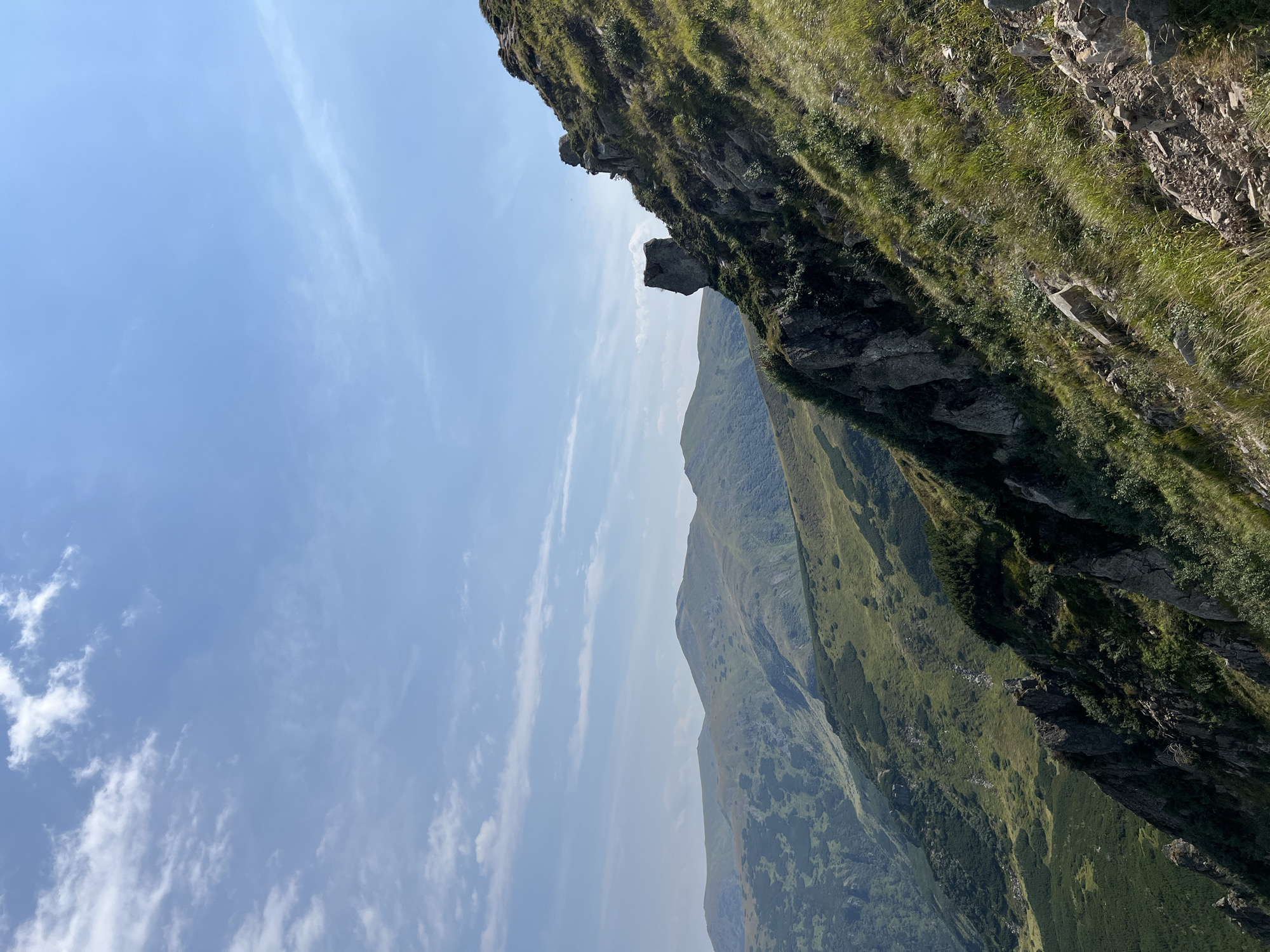 The image depicts a mountainous landscape under a partly cloudy blue sky. In the foreground, a steep, grassy slope is dotted with rocks and some vegetation. Further down the slope, sharp rock formations are visible. In the middle ground, a prominent, uniquely shaped rock formation stands out on the side of a mountain. Beyond that, rolling green mountains stretch into the distance, with the terrain showing variations in vegetation and texture. The overall scene suggests a natural, outdoor environment in a mountainous region. The provided location of Vorokhta, Ukraine, implies this is a view of the Carpathian Mountains. There are no people or specific man-made objects visible in the image, focusing solely on the natural topography and sky.