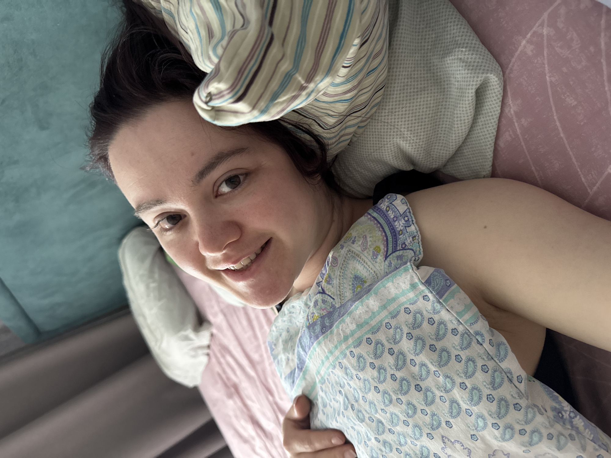 A woman with dark hair is lying in bed. She is smiling at the camera. She is holding a white and blue patterned blanket. There are pillows behind her head, one striped and one light green. The bed has a pink sheet. The location is not discernible from the image. There is no evidence of Izhevsk, Russia.