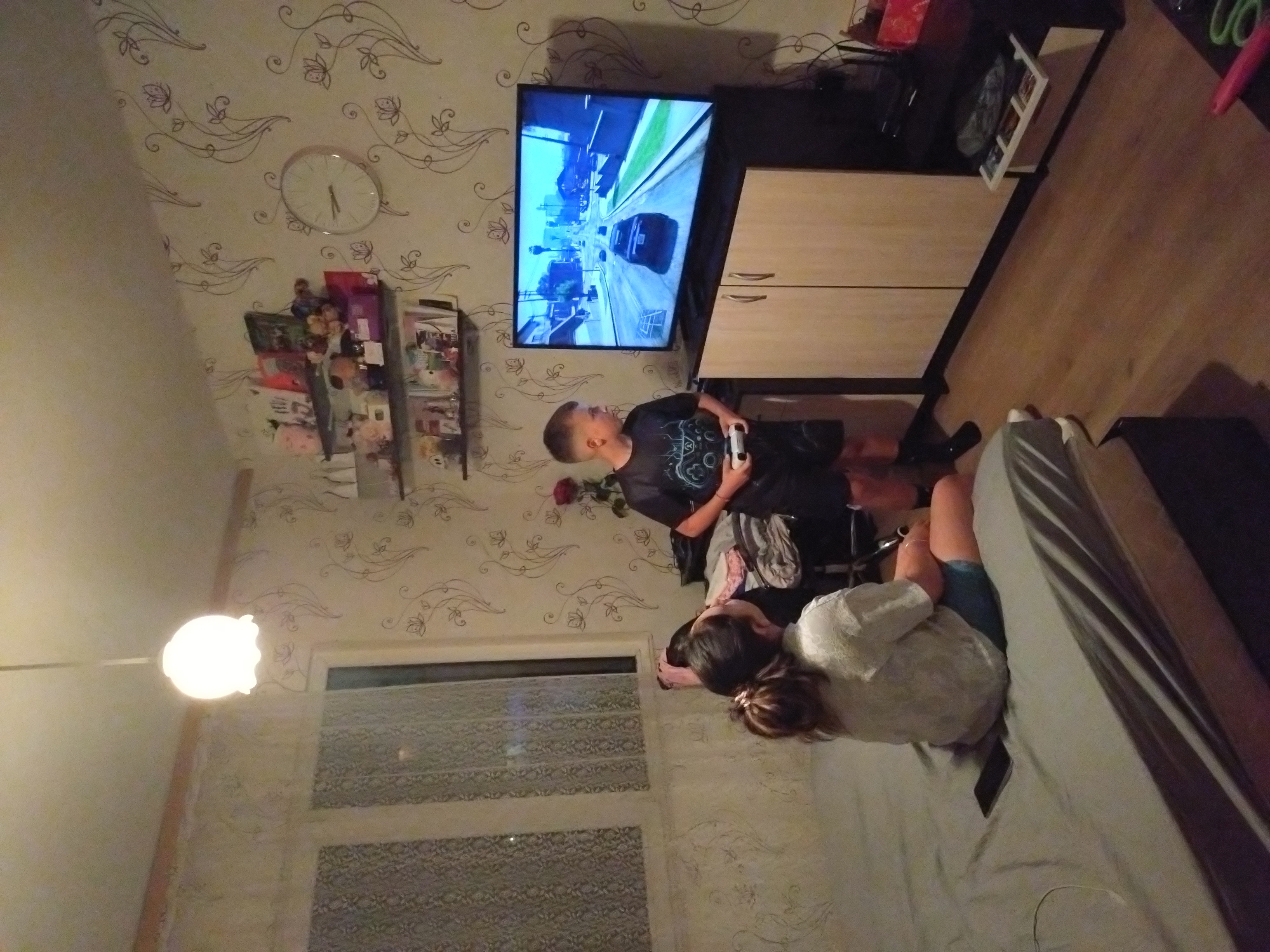 A slightly distorted, high-angle indoor photograph shows a boy playing a video game, with another person lying on a bed in the foreground.
The boy is standing in front of a flat-screen TV, holding a white game controller. The TV is displaying a video game with a cityscape and a car on the screen. Beside the TV is a light-colored cabinet. To the left of the TV, on the wall, are a clock and various items on shelves.
In the foreground, a person is lying on a bed. The bed has striped bedding.
The room has patterned wallpaper and a window with patterned blinds. An overhead light fixture is visible.
The image gives no explicit information that places this at Stara Zagora, Bulgaria.