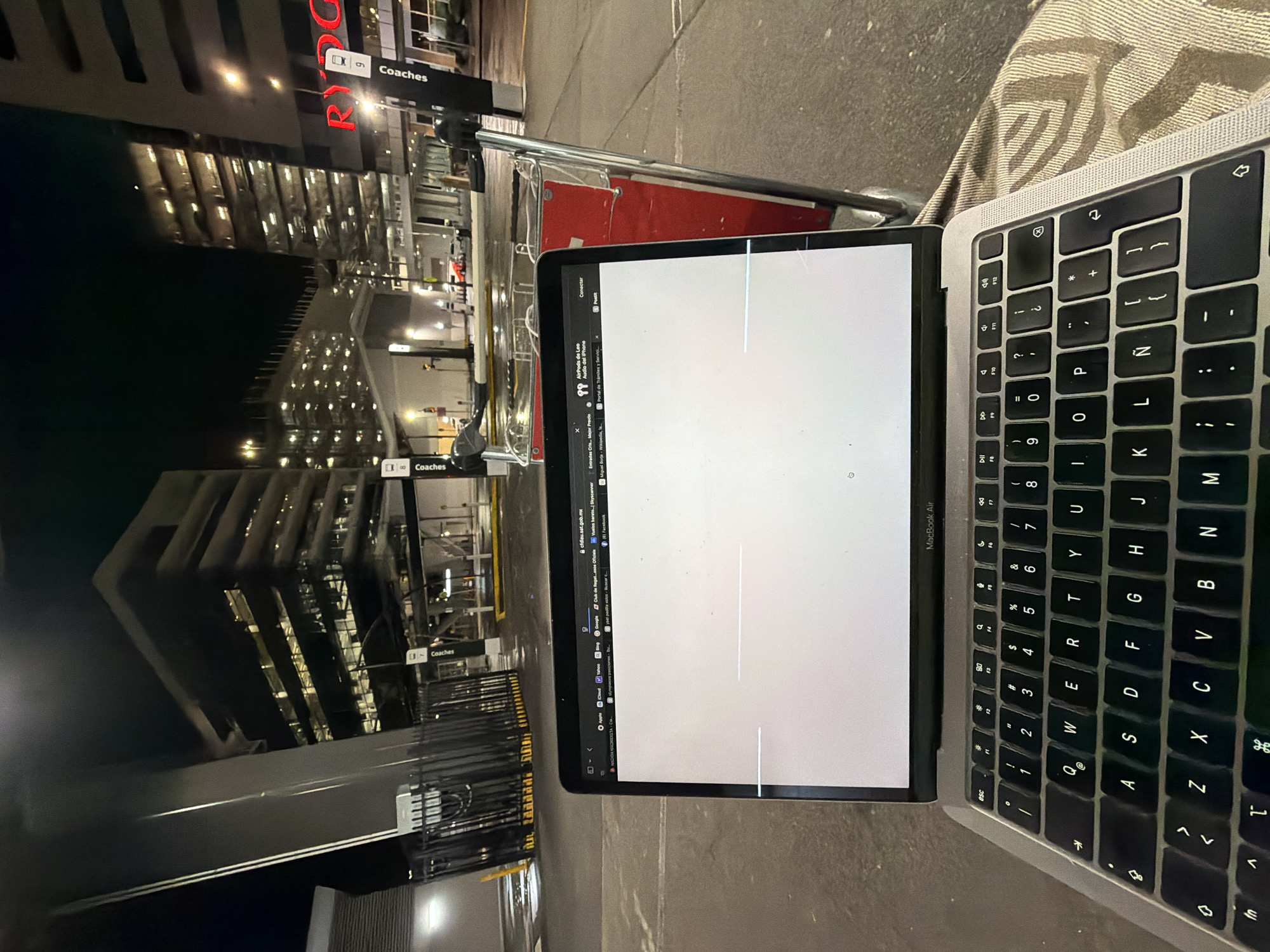 The image captures an outdoor scene at night, likely an airport terminal or transport hub, given the presence of a luggage trolley and modern building architecture. The foreground is dominated by an open laptop, a MacBook Air, placed on a textured fabric surface. The laptop screen displays a blank white page with faint horizontal lines. The keyboard is visible, showing a standard QWERTY layout with Spanish characters.
Behind the laptop, a red and silver luggage trolley is positioned, its metallic frame reflecting the ambient light. In the background, multi-story buildings with illuminated windows suggest a commercial or transport-related complex. The architecture is modern, characterized by glass facades and structured elements. Road markings and traffic lights are discernible on the wet asphalt, indicating a vehicle-accessible area. Signs with the word "Coaches" are visible, suggesting a bus or coach terminal. The overall atmosphere is urban and functional, illuminated by artificial lighting that casts a slightly eerie glow on the scene. There are no people visible in the image. The location is identified as Mascot, Australia.