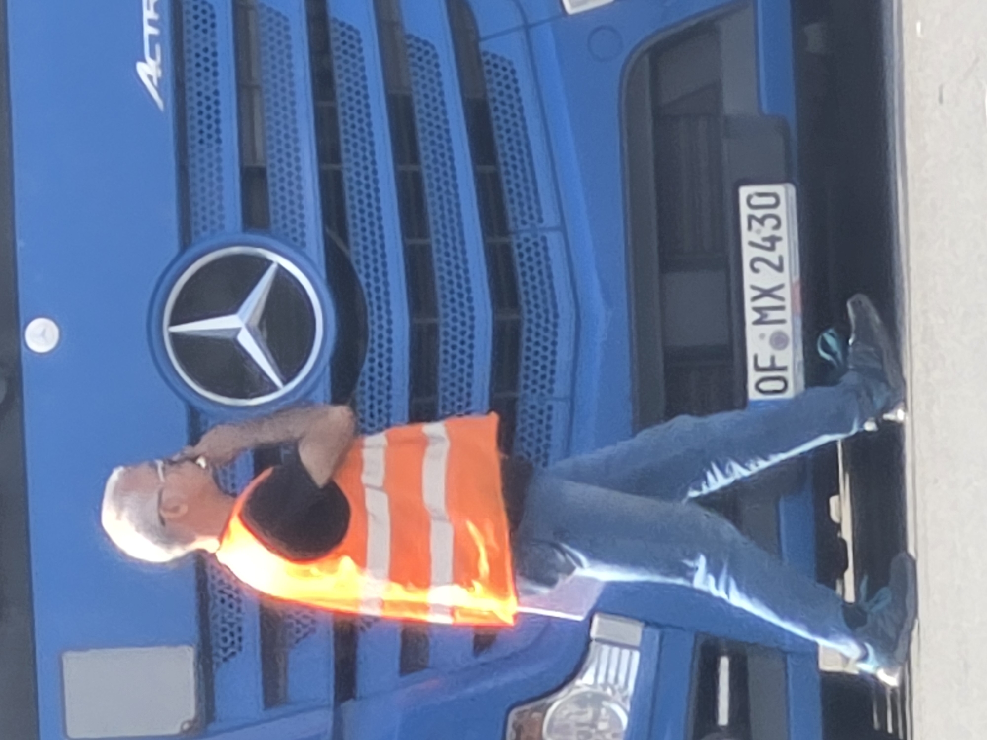 A man with gray hair and glasses is walking in front of a blue Mercedes-Benz Actros truck. He is wearing a black t-shirt, an orange high-visibility vest, and blue jeans. The truck has a Mercedes-Benz star logo on the grille. A license plate on the truck reads "F-MX 2430". The scene is outdoors, with a bright, overcast sky. The location is identified as Frankfurt, Germany, by the license plate.