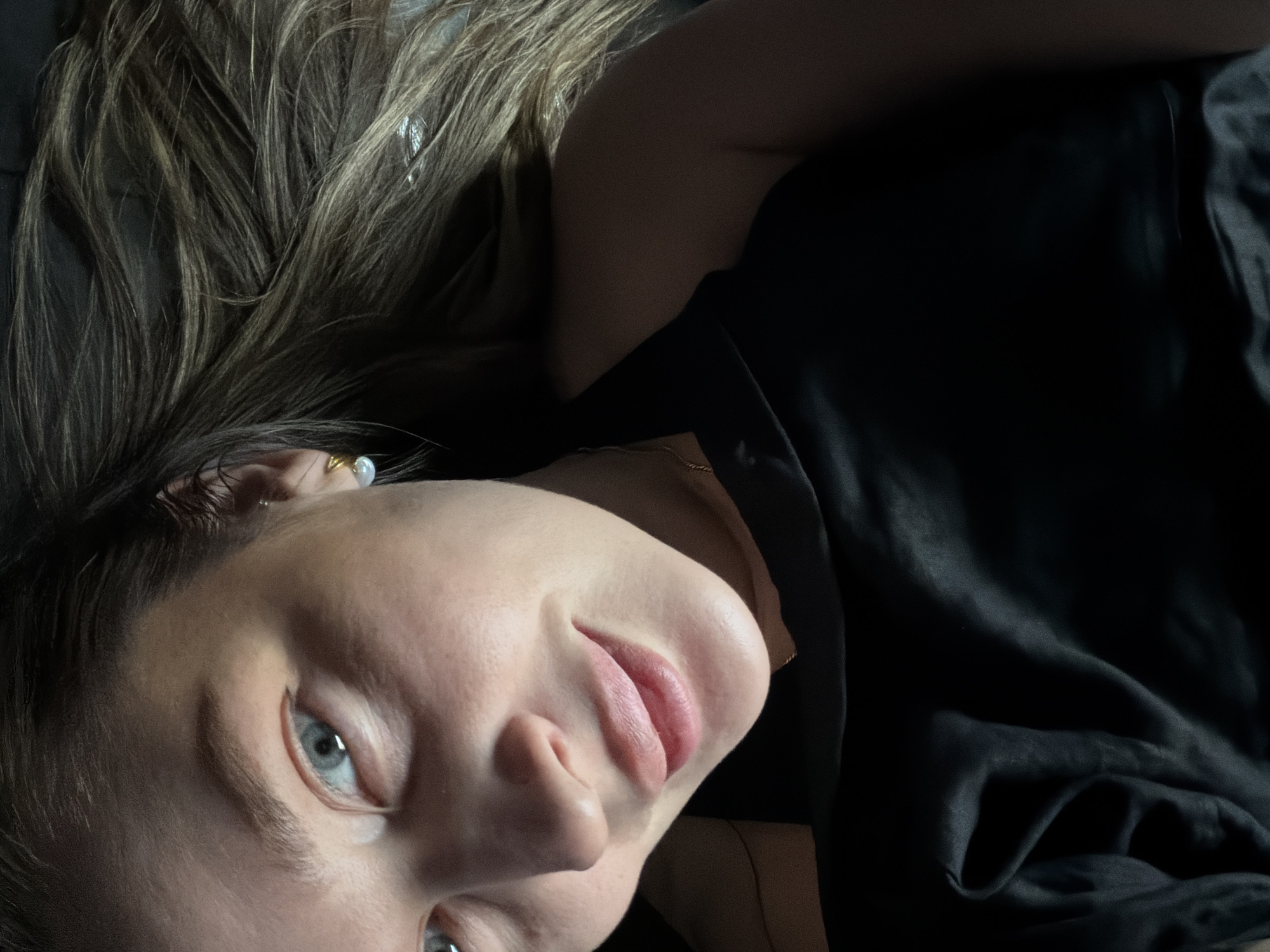 A woman lies on a dark surface, possibly a bed, with her head tilted towards the camera. She has fair skin, blue eyes, and light brown hair that is spread out above her head. She wears a black top, and a small pearl earring is visible on her left ear. A thin gold chain is visible around her neck. The background is indistinct and dark. No information indicates that this image was taken in Moscow, Russia.