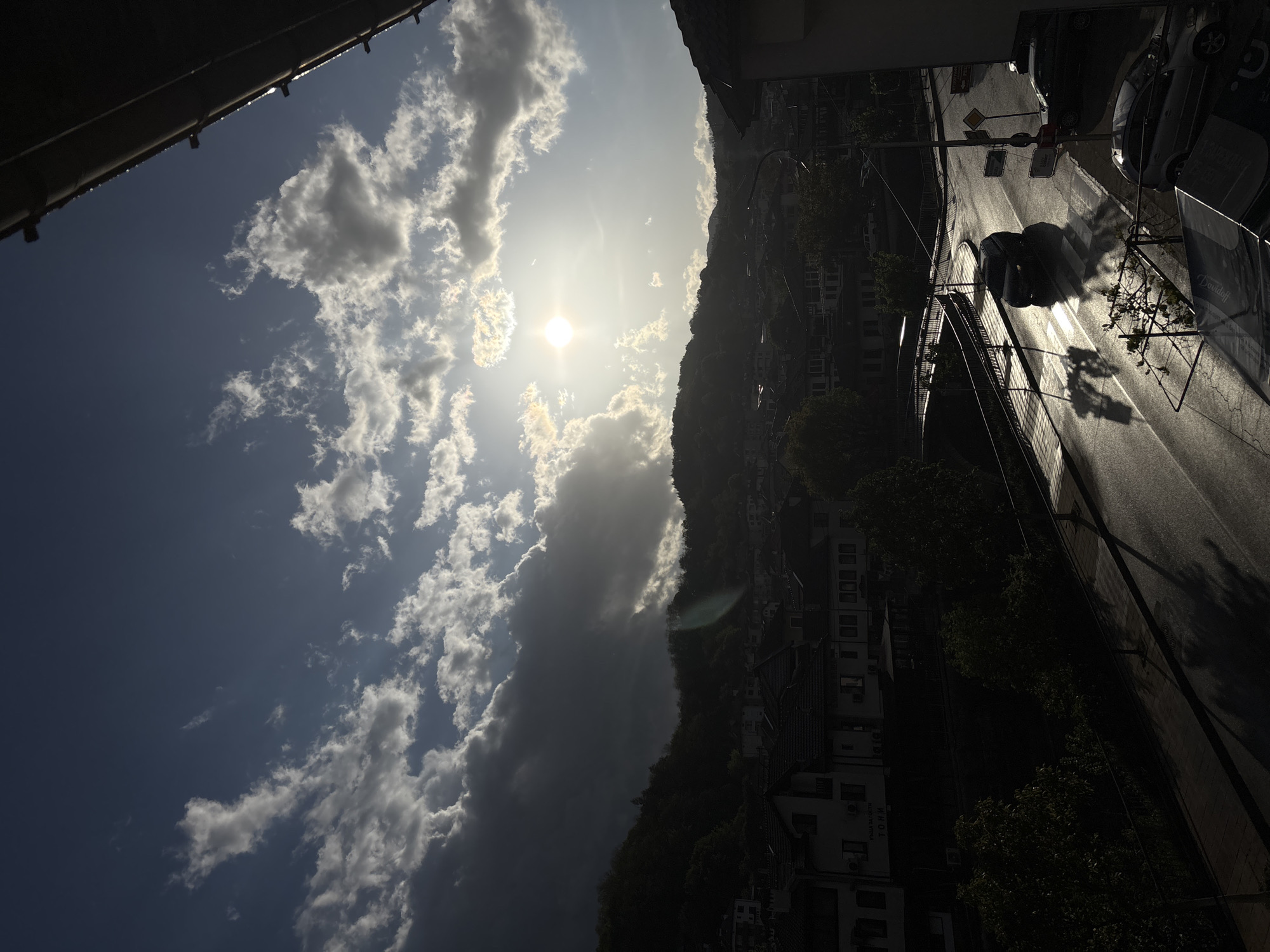 The image shows a view from above, likely a high vantage point like a building. The upper left corner shows a partial view of a building's structure. The sky occupies a significant portion, displaying a mix of blue sky and white and gray clouds, with the sun shining brightly through them.
To the right of the clouds, a hillside covered in buildings and trees descends. This landscape is likely Zlatograd, Bulgaria, given the urban layout nestled within a hilly or mountainous terrain.
On the right side of the image, a road is visible, reflecting sunlight. Cars are parked along the side of the road. There are also shadows of trees and structures cast onto the road surface. Traffic lights are also present, suspended over the street. There are street signs visible as well.