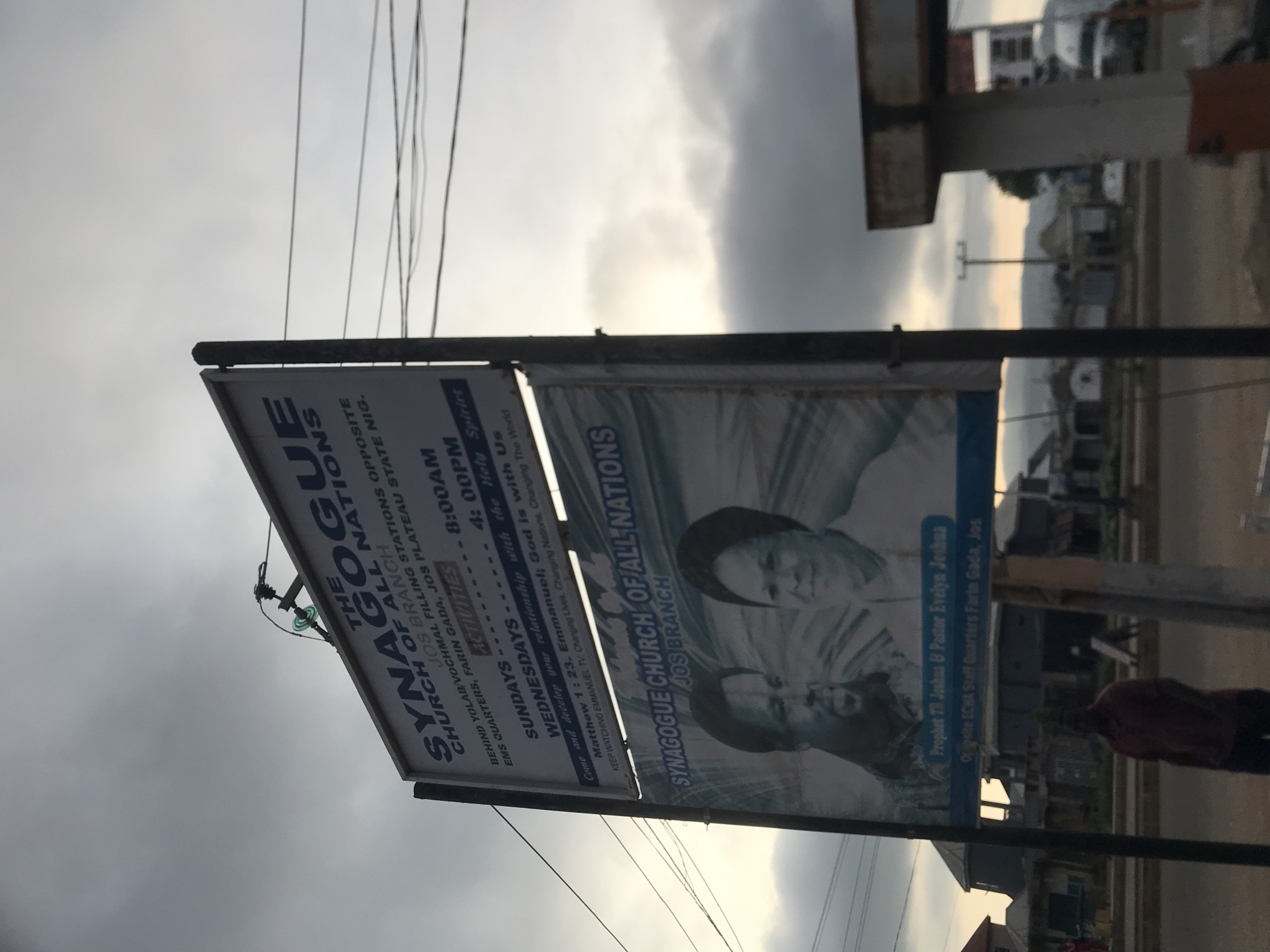 A vertically oriented image depicts a large advertising billboard for "The Synagogue Church Of All Nations Jos Branch" set against a cloudy, overcast sky. Multiple electrical wires and cables crisscross the upper part of the frame.
The billboard features two main sections. The left section, mostly white with dark blue text, provides the church's address: "BEHIND YOLAB/VOCHMAL FILLING STATIONS OPPOSITE EMS QUARTERS, FARIN GADA, JOS PLATEAU STATE NIG." It lists service times as "SUNDAYS - 8:00AM" and "WEDNESDAYS - 4:00PM" under "ACTIVITIES." Further text includes spiritual messages: "Come and develop your relationship with the Holy Spirit," "Matthew 1:23. Emmanuel; God is with Us," and an encouragement to "KEEP WATCHING EMMANUEL TV, Changing Lives, Changing Nations, Changing The World."
The right section of the billboard displays two grayscale portraits, identified by accompanying text as "Prophet TB Joshua & Pastor Evelyn Joshua." This section also repeats "SYNAGOGUE CHURCH OF ALL NATIONS JOS BRANCH" and adds "Opposite ECWA Staff Quarters Farin Gada, Jos."
In the background, a street or open area is visible with distant buildings and what appear to be hills or a hazy horizon under the heavy cloud cover. The muted lighting suggests either an early morning, late afternoon, or a very overcast day. A person in a dark shirt is partially visible on the right side, next to the billboard. The scene is an outdoor setting in Rafin Par, Nigeria, characteristic of a semi-urban or suburban environment. No specific activity is taking place beyond the presence of the advertisement.