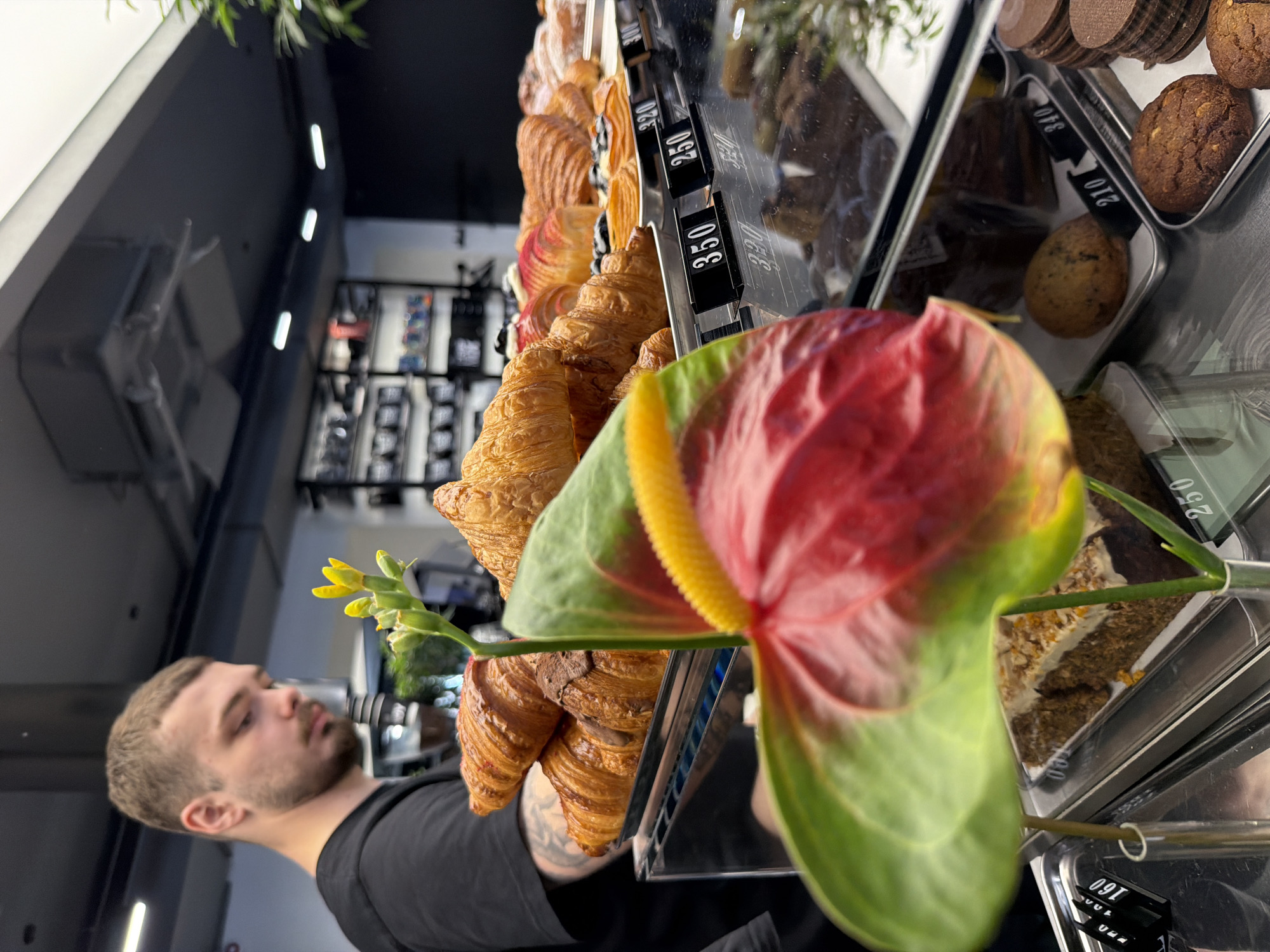 The image presents a close-up view of a bakery display cabinet, likely situated in Izhevsk, Russia, given the Cyrillic script visible on the price tags.
The cabinet is filled with various pastries, primarily croissants in the foreground. Their price tags, in Cyrillic lettering, are attached to the front of the display. Cookies and other baked goods are visible in the background compartments.
Foreground elements include a man partially visible on the left, seemingly near the display cabinet. He wears a black t-shirt. Interspersed with the croissants are flowers, namely a red and green anthurium and a stem of small yellow flowers, placed decoratively within the display.
The environment suggests a modern bakery or café, indicated by the lighting, the glass display case, and the pastries. The setting includes elements like shelves of items and lighting.