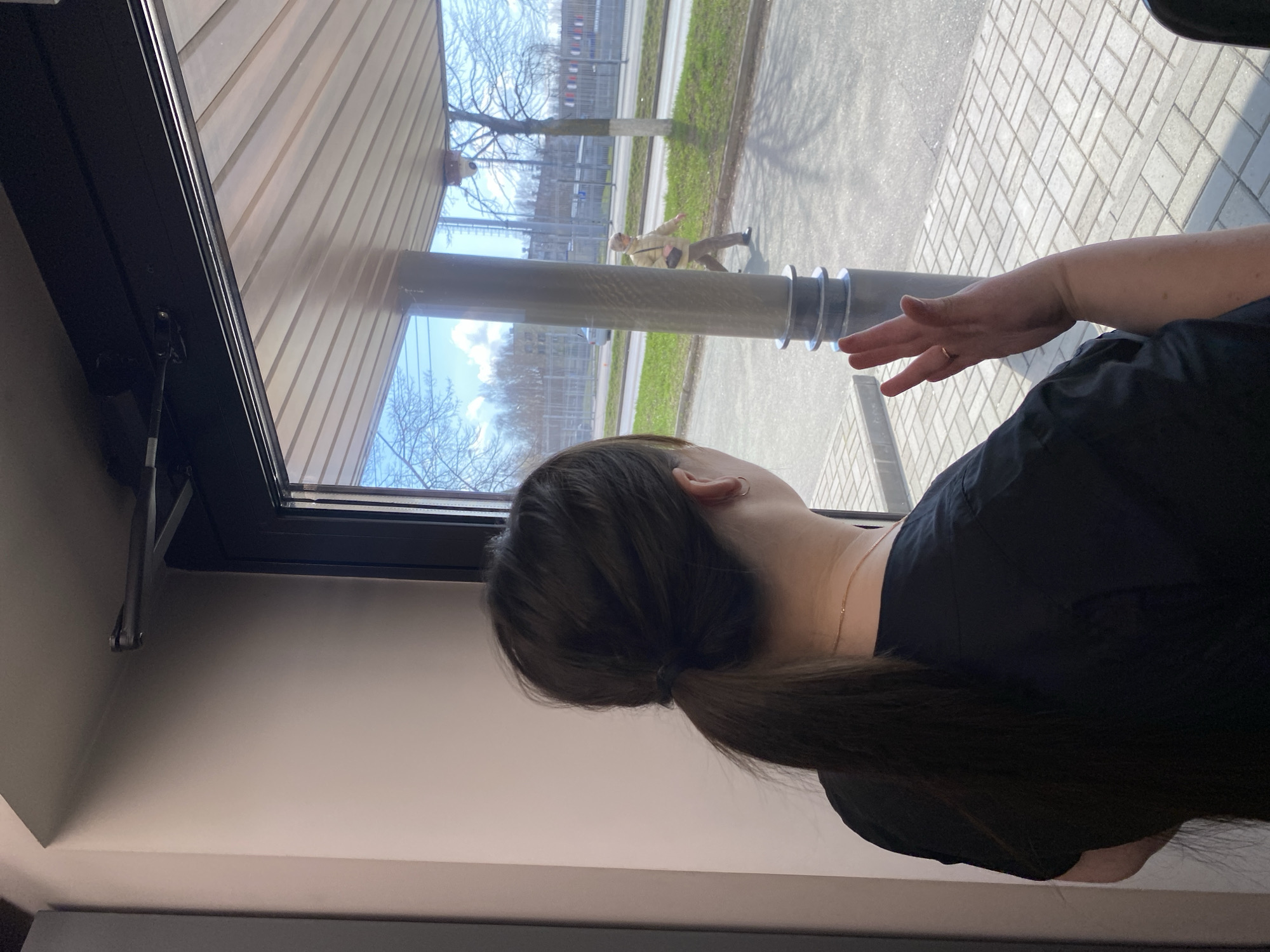 A woman is standing indoors, looking out of a window. Her dark hair is pulled back in a ponytail. She is wearing a black garment. Her right hand is raised, fingers extended, near the window. Through the window is a scene in Votkinsk, Russia: a person walks along a sidewalk bordering a green, grassy area. Across the street is a fenced structure, possibly an industrial or commercial building. The sky is visible through bare tree branches.