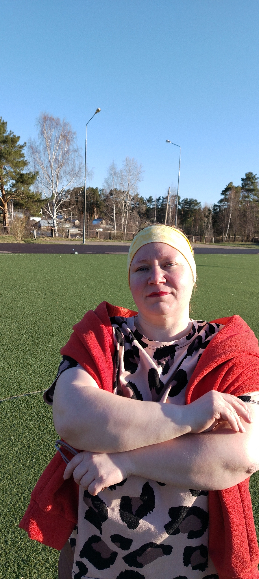 The image depicts a woman standing on a green artificial turf field. She is wearing a yellow headband and a t-shirt with a leopard print pattern. A red garment is draped over her shoulders and her arms are crossed.
In the background is a row of trees and a few houses. There are also two streetlights visible. The sky is blue and clear.
Based on the location information, this image was taken in Chaikovsky, Russia.