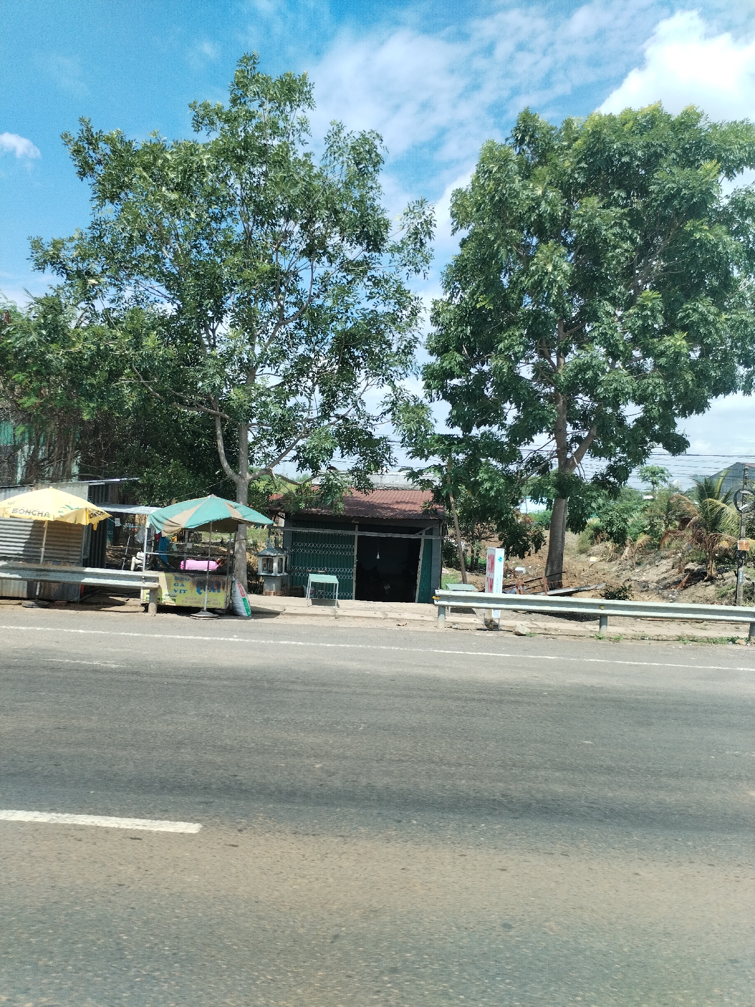 The image captures a street scene in Ia Le, Vietnam. A two-lane asphalt road with a white dividing line takes up the foreground. Beyond the road, there is a roadside area featuring a few small businesses. On the left, there is a yellow and white umbrella, presumably shading a vendor's stall. Adjacent to it, another stall is covered by a green and white striped umbrella. In the background, a small, single-story building with a red roof can be seen. The building has green corrugated metal walls with a partially open storefront. To the right of the building, there are two large trees. A metal guardrail stretches along the edge of the road, separating it from the roadside area and a sloped embankment covered in vegetation. The sky is blue with scattered clouds.