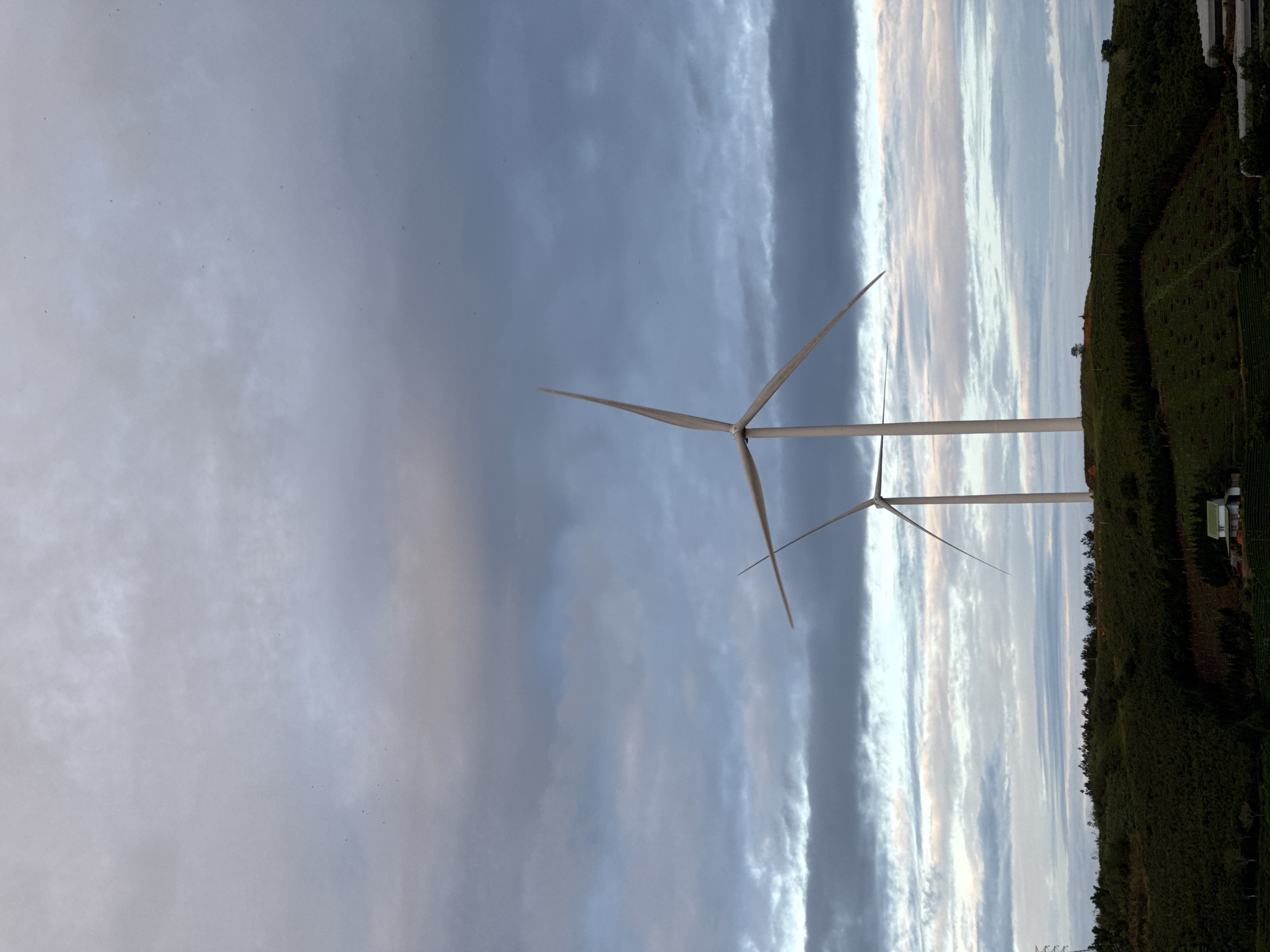 The image shows two wind turbines standing on a hillside in Phát Chi, Vietnam. The turbines are white and have three blades each. The sky is cloudy with shades of gray and light blue. The hillside is covered in green vegetation.