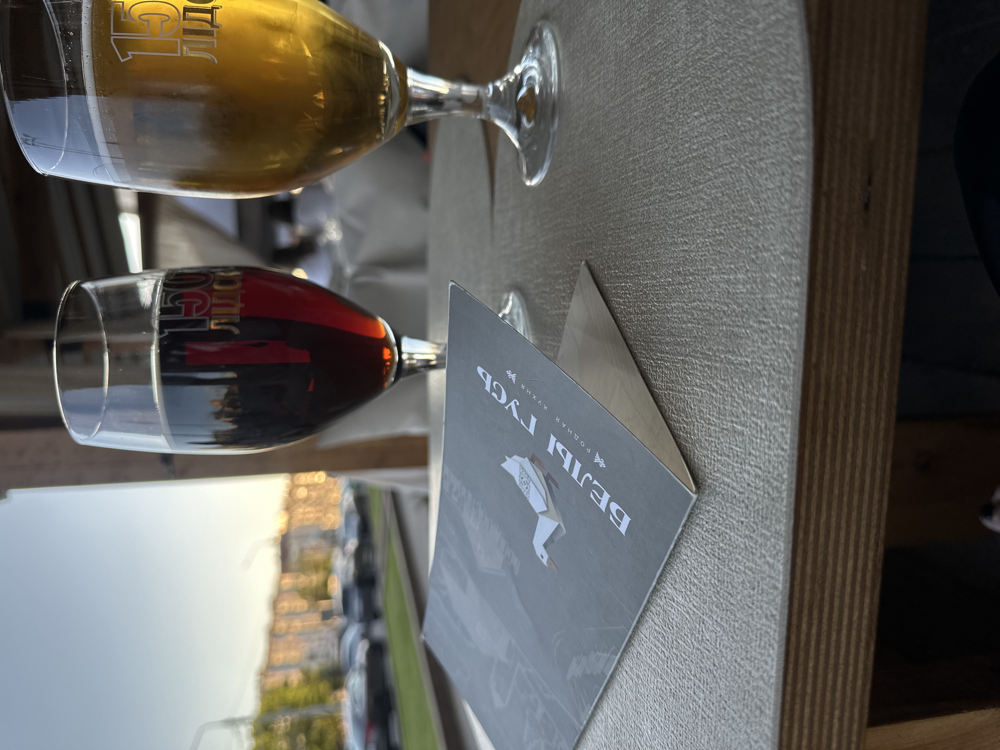 Two glasses of beer, one dark and one light, sit on a textured table next to a menu with a white origami goose and the Cyrillic text "БЕЛЫ ГУСЬ" and "РОДНАЯ КУХНЯ". In the background, a blurred city scene includes buildings and cars under a clear sky. The lighting suggests it is late afternoon or early evening. This scene appears to be at an outdoor seating area of a restaurant, possibly overlooking a street. The establishment's name "Белый гусь" (White Goose) and "Родная кухня" (Native Cuisine) are visible on the menu, suggesting a Belarusian restaurant. Based on the visible text, the location is likely Minsk, Belarus, where this restaurant chain has a presence.