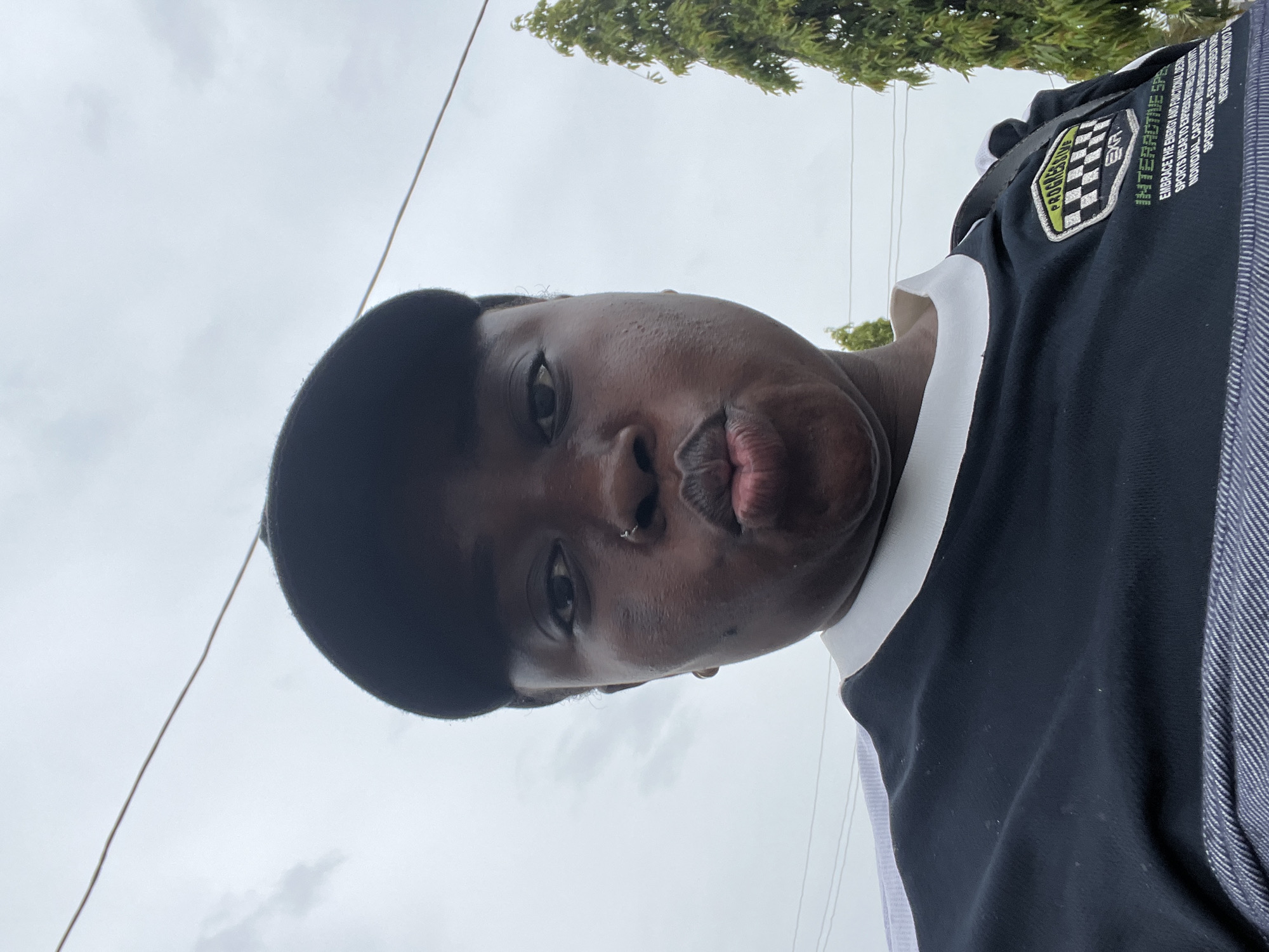 A person wearing a black baseball cap and a black and white collared shirt is posing for a selfie outdoors. The person has a nose ring and is puckering their lips. In the background, there is a cloudy sky, power lines, and a tall green tree. The scene appears to be in an urban or suburban setting in Abuja, Nigeria.