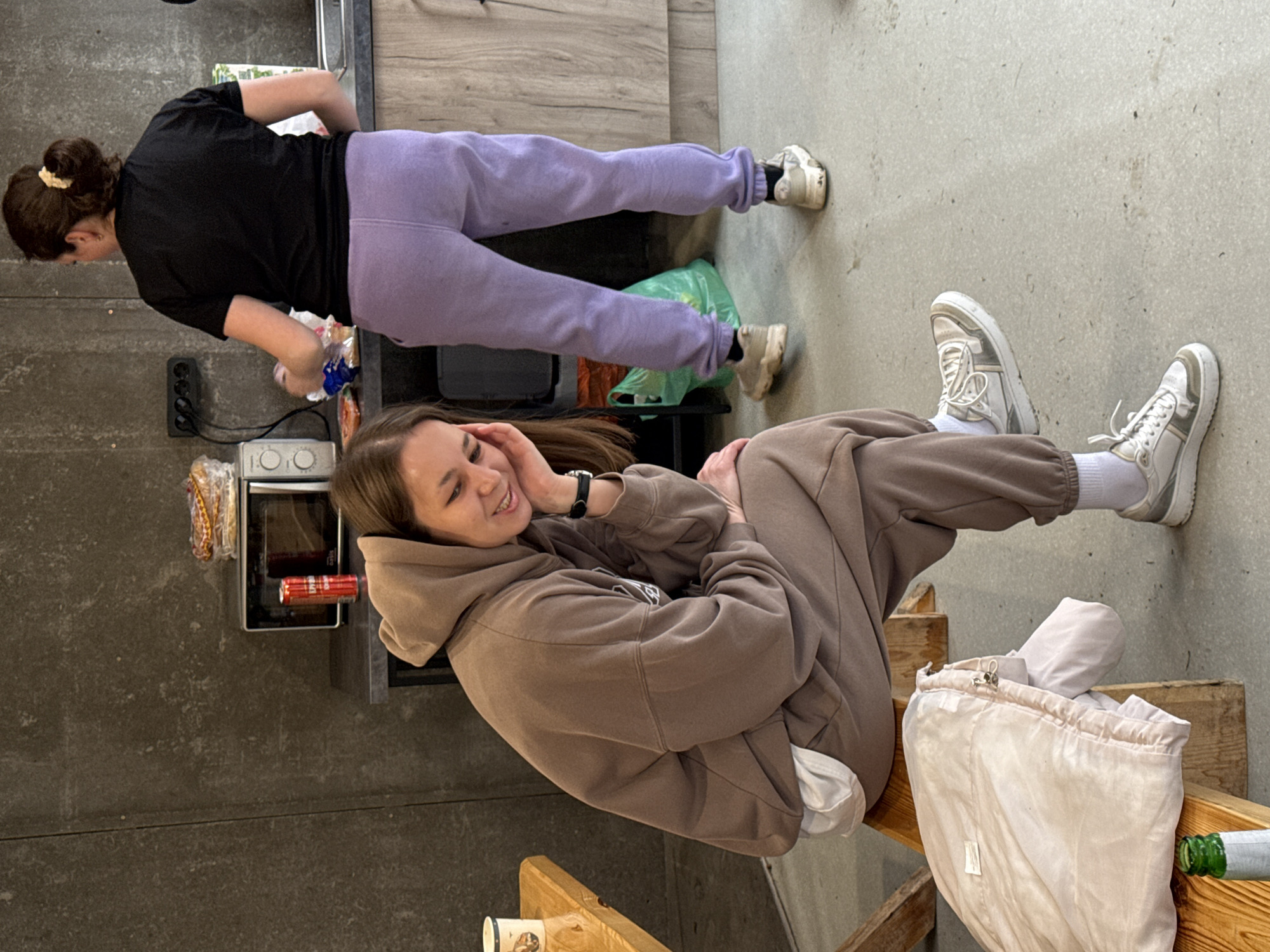 Two young women are seen in a casual indoor setting, likely a home or communal space, in Yekaterinburg, Russia. The image is taken from a slightly elevated angle, looking down.
In the background, a woman with dark brown hair tied in a bun with a light scrunchie is standing. She is wearing a black short-sleeved top and light purple sweatpants, paired with light-colored chunky sneakers. She appears to be leaning over a light wooden counter or cabinet, possibly attending to something in a kitchen area. Near her feet, a green plastic bag and another orange/brown bag are visible on the grey floor. A microwave and a smaller appliance (possibly a toaster oven) are on a counter to her right, next to a black electrical outlet with cables.
In the foreground, another woman with long brown hair is seated on a wooden structure, possibly a bench or steps. She is wearing a brown hooded sweatshirt and matching sweatpants, with white socks and white and grey sneakers. She is resting her chin on her hand, smiling, and looking towards the left side of the frame. A black watch is visible on her left wrist. Next to her, on the wooden structure, is a light beige drawstring bag with a metal clasp, and a green glass bottle. A white disposable cup with a pattern is also on the wooden surface.
The floor is a textured grey, possibly concrete, and a darker grey wall is seen in the background. The overall impression is one of a relaxed, informal gathering. No distinct text is clearly visible on any objects.
