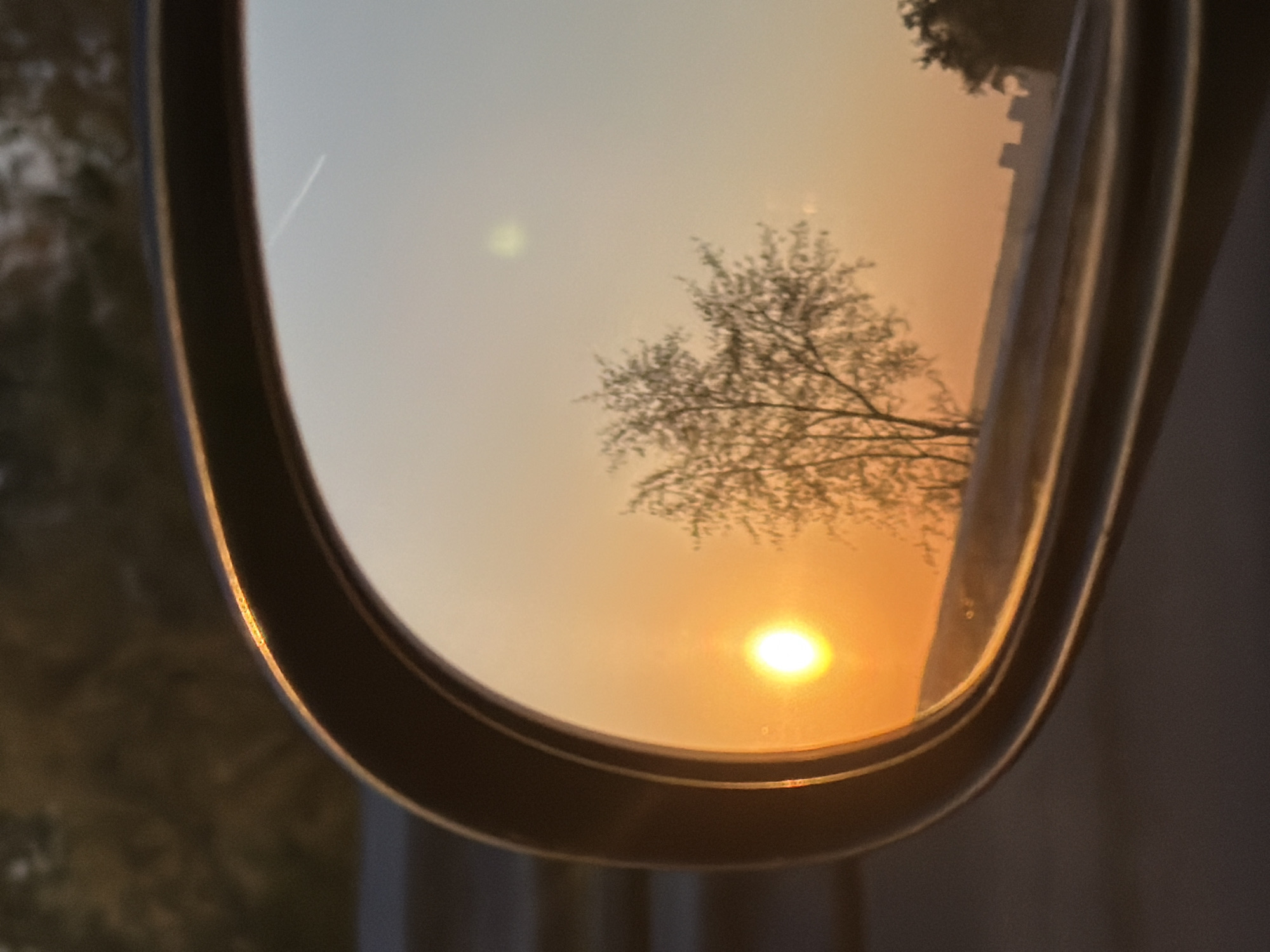 The image shows a sunset reflected in a car's side mirror. A bare tree is visible in the reflection, silhouetted against the orange and yellow sky. The sun is a bright spot near the bottom of the mirror. The location and details related to Izhevsk, Russia, are not deducible from the provided image.