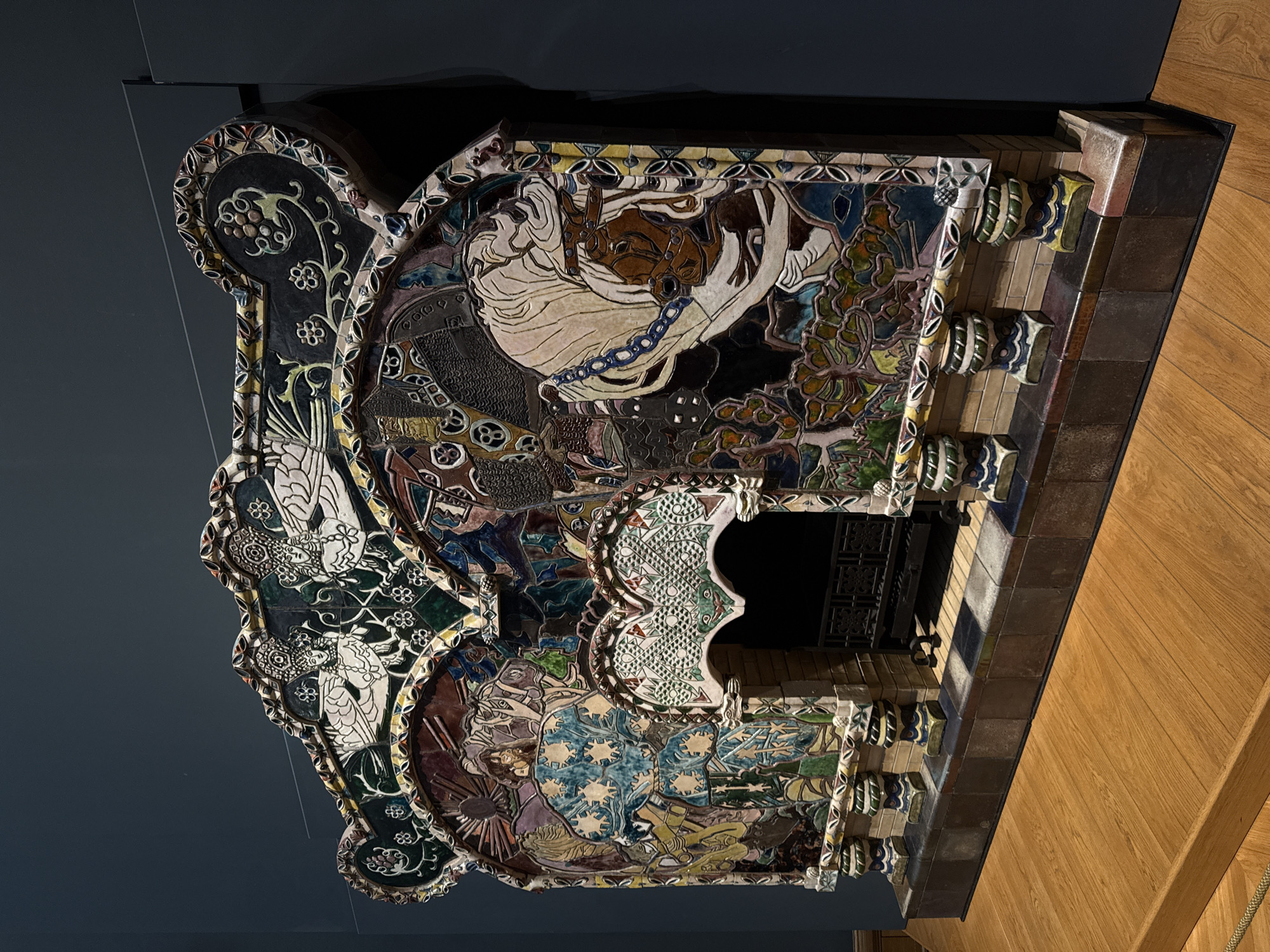 An elaborately decorated tile fireplace is displayed against a dark blue wall and wood flooring. The fireplace features intricate, colorful tilework depicting figures, patterns, and foliage. The scene is cropped, offering an unusual perspective, possibly taken in a museum or art gallery. No people are visible. The city and country are not discernable from this information.