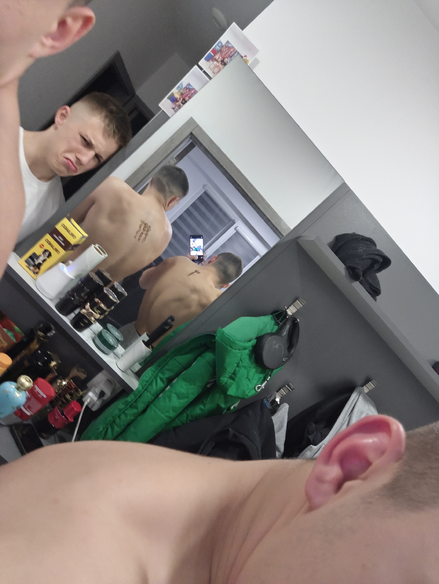 The image is a selfie in a bathroom with a mirror. A young man with short, light brown hair is visible in the reflection. He has a slight frown. He is wearing a white t-shirt. His bare back is also visible in the reflection and appears to have a small tattoo. He is holding a smartphone, possibly taking the selfie. In the mirror's reflection, there is a window with blinds. The bathroom countertop is cluttered with various items, including perfume bottles and boxes. A green jacket and a black bag hang from hooks on a gray cabinet to the right of the mirror. There is no discernible information to determine the location is in Kozloduy, Bulgaria.