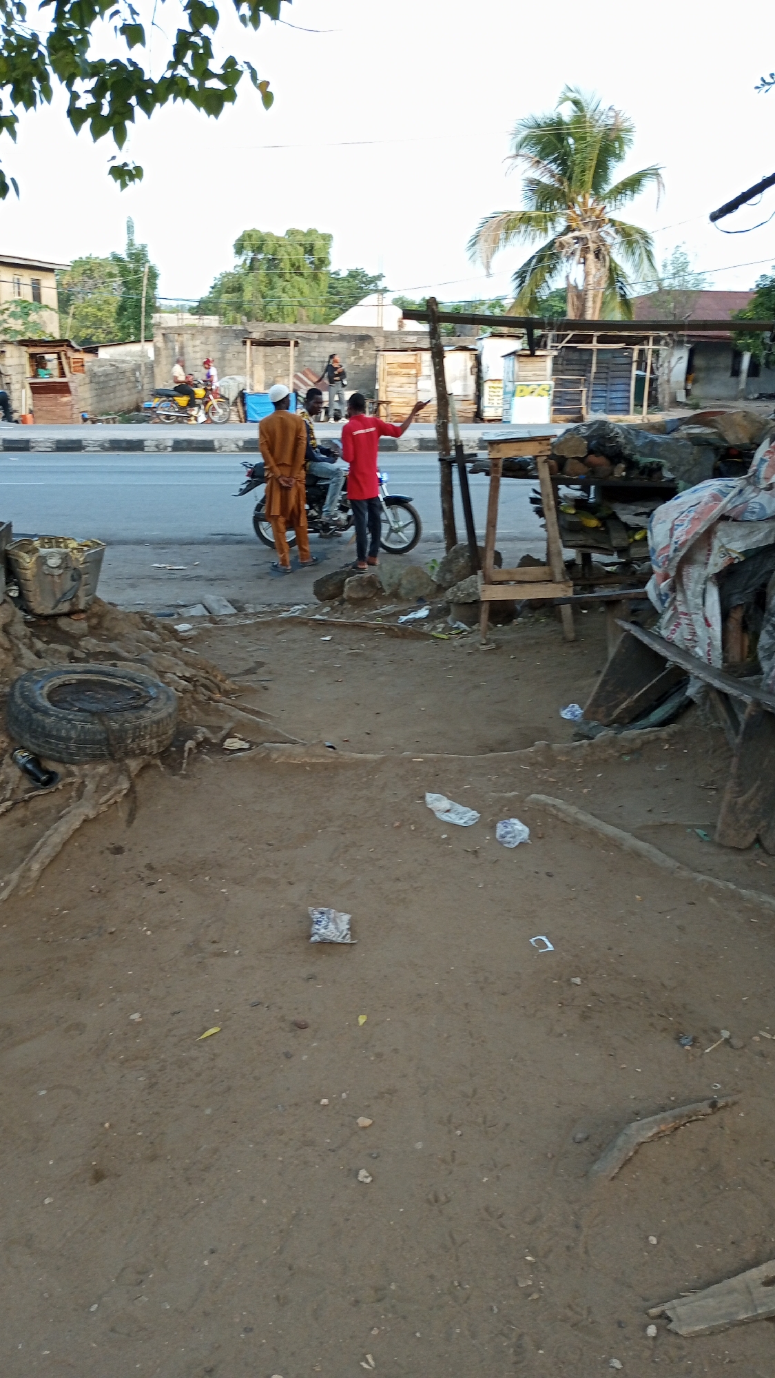 The image captures a street scene in Adankolo, Nigeria. In the background, several people are visible on the street, including two men on a motorcycle and another man in a red shirt gesturing with his hand. There are buildings and informal stalls or shops lining the street, and a palm tree is visible in the distance. The foreground shows a dirt or sandy area with some debris, including a tire, pieces of wood, and scattered plastic. The scene appears to be a typical street view in a busy urban area.