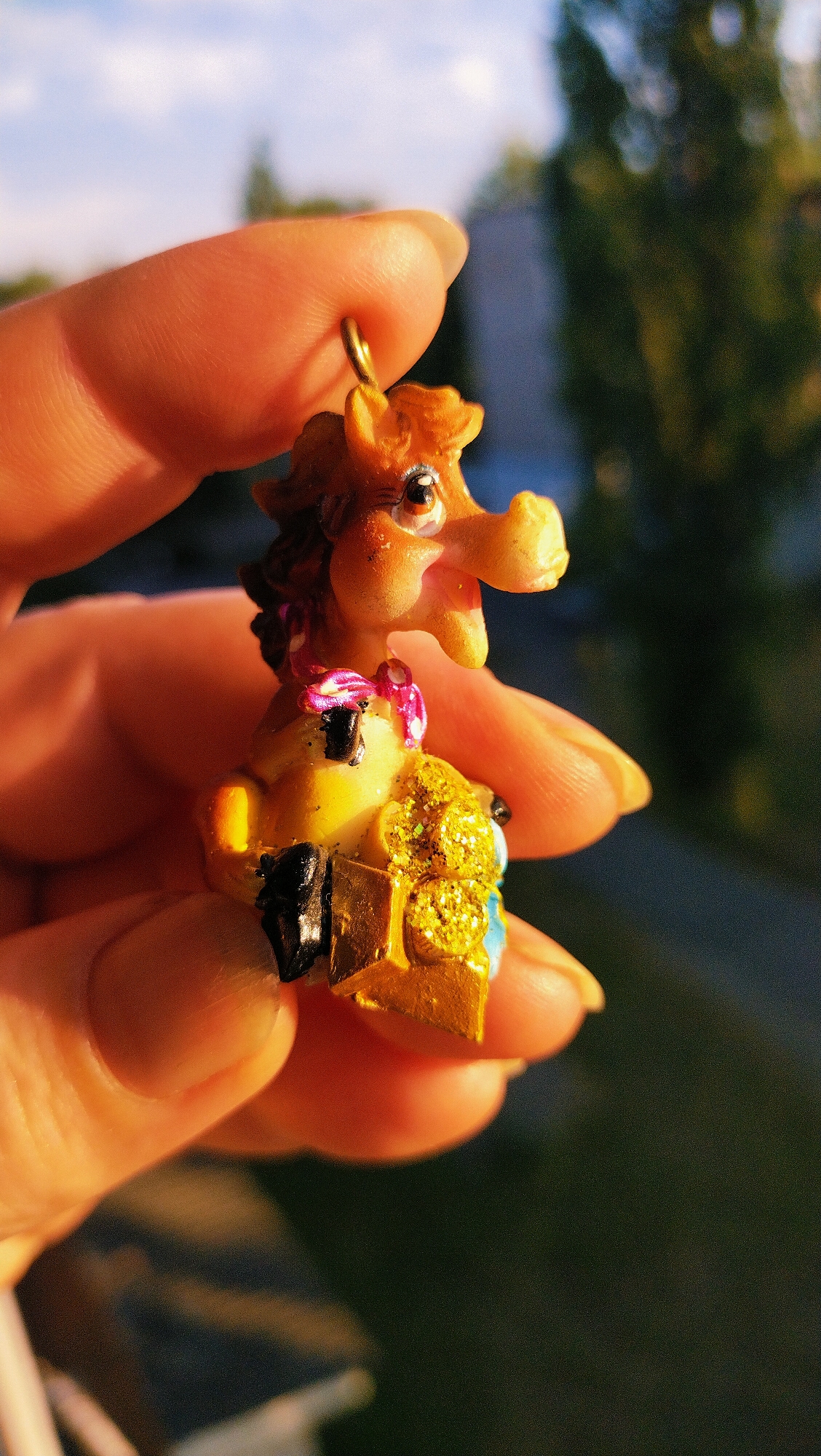 A close-up, high-angle, eye-level outdoor shot features a person's hand holding a small, brown, horse-shaped figurine, possibly a charm or pendant. The figurine has a brown mane, large eyes with black pupils and white highlights, and a light brown snout with an open mouth. It is adorned with a pink ribbon tied around its neck and sits on a golden, glitter-covered object. The background is blurred, showing a green, leafy tree and a bright blue sky with white clouds, indicative of a sunny day. The lighting suggests it is either sunrise or sunset, casting a warm glow on the scene, which may be located in Horishni Plavni, Ukraine.