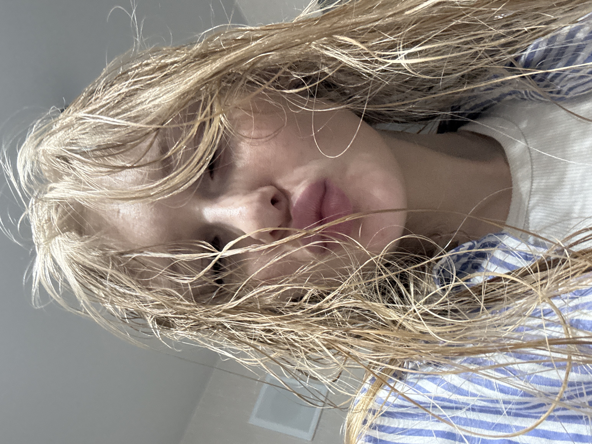 A close-up, upside-down selfie photograph depicts a woman with wet, blonde hair obscuring much of her face. Her eyes are partially closed, and her lips are visible. She is wearing a white shirt underneath a blue and white striped shirt. The background shows a white wall with a rectangular feature, possibly an air vent. There is no indication of the location being Perm, Russia, or any specific city.