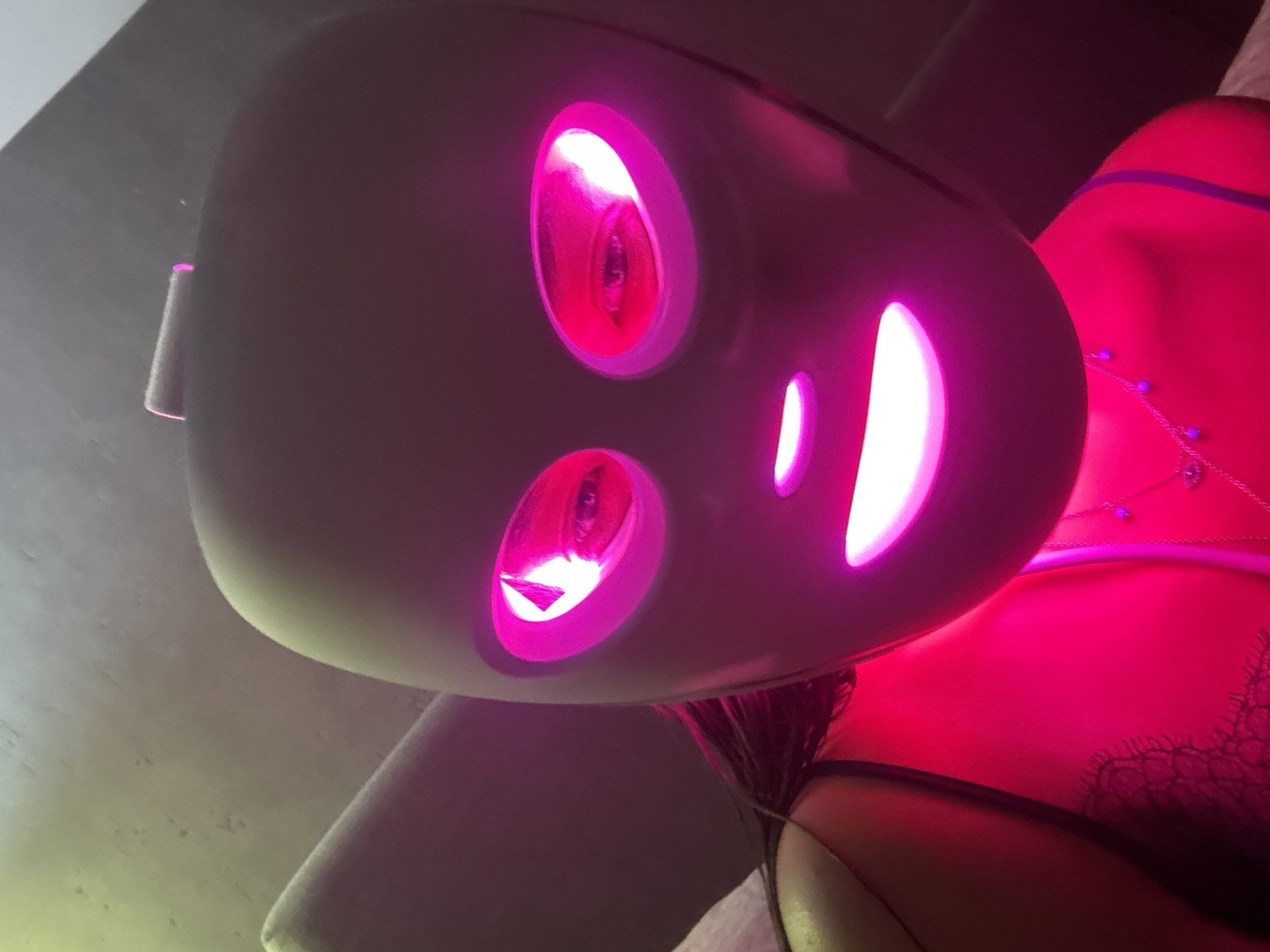 The image presents a close-up, low-angle perspective of a person wearing a dark, futuristic-looking mask that emits a vibrant pink light. The mask features illuminated cutouts, creating glowing eyes and a distinct smiling mouth. Beneath the mask, a person's upper body is visible, adorned in what appears to be bright pink or red clothing, possibly lingerie, with intricate black lace detailing along the edges. A delicate chain necklace, embellished with small purple beads and a central charm, rests on the person's chest. Dark hair is also visible peeking out from below the mask. The intense, artificial pink illumination from the mask and the clothing dominates the color palette, suggesting an indoor setting, likely in dim ambient light to enhance the glow. There is no visible text within the image. The scene is located in San Bernardino Tlaxcalancingo, Mexico.