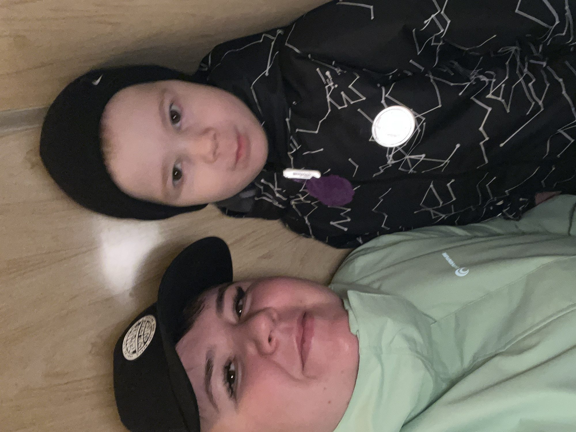 An adult female and a young child are depicted in a close-up portrait, likely a selfie, within the city of Chaikovsky, Russia. The background is a uniform, light brown surface, suggesting an indoor wall or paneling with vertical seams.
The adult female, positioned on the left, wears a black baseball cap with a white circular logo and a light mint green hooded jacket bearing a white "Outventure" logo on the chest. She has dark hair, prominent eyelashes, and displays a slight smile while looking directly at the camera.
The young child, positioned to the right, wears a black beanie with a white Nike swoosh logo and a black hooded jacket patterned with white abstract geometric or constellation-like lines. A purple pom-pom ornament is attached to a zipper pull on the jacket's left side, and a round, silver-colored reflective badge is affixed to the right side of the jacket. The child has reddish-brown hair visible under the beanie and maintains a neutral expression, looking directly forward.